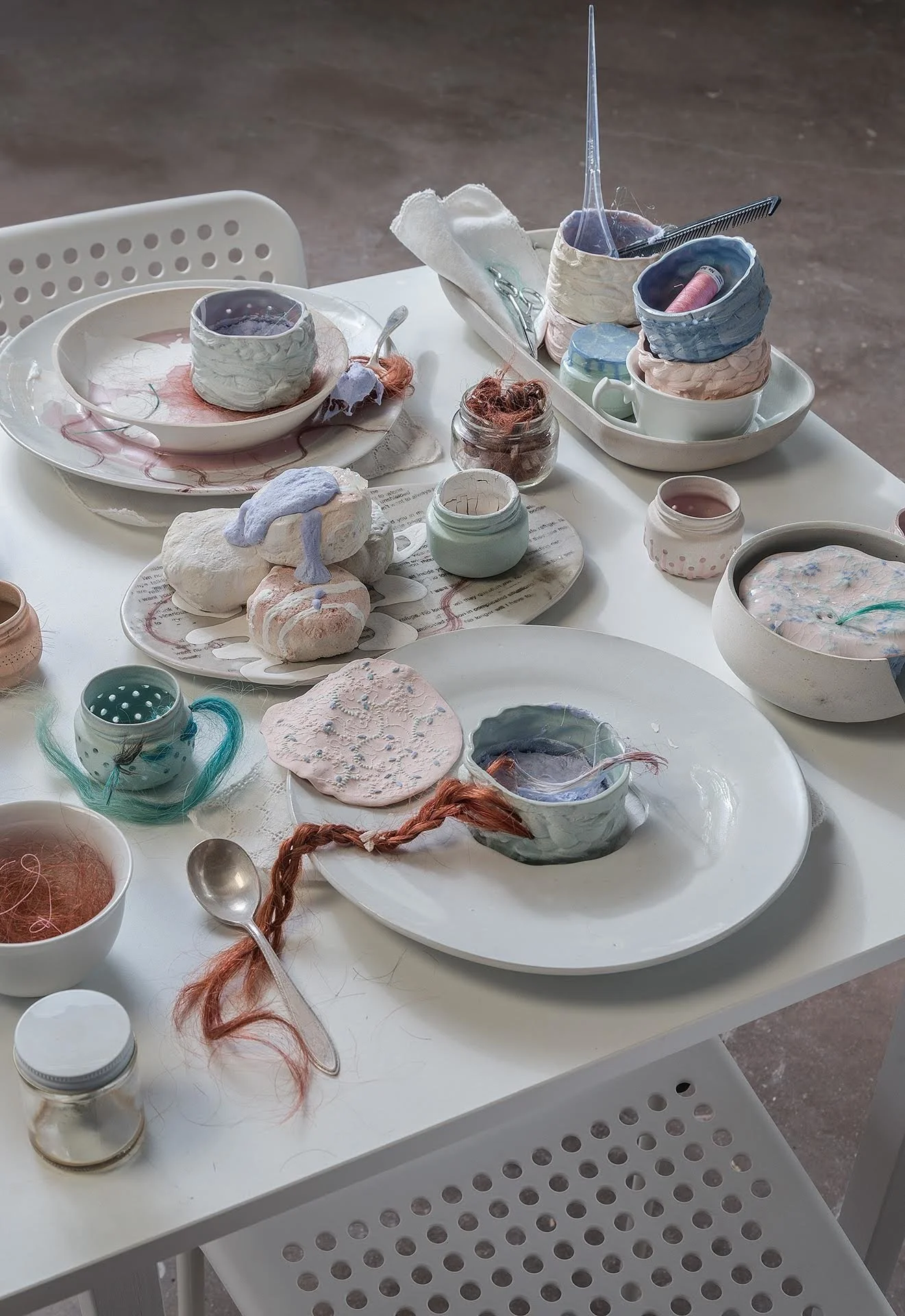 Tabletop cluttered with dishes that have red hair woven through; ceramic biscuits in the center of the table covered with foamy purple bleach. Jars and assorted cups cast with the pattern of braided hair fill the space.