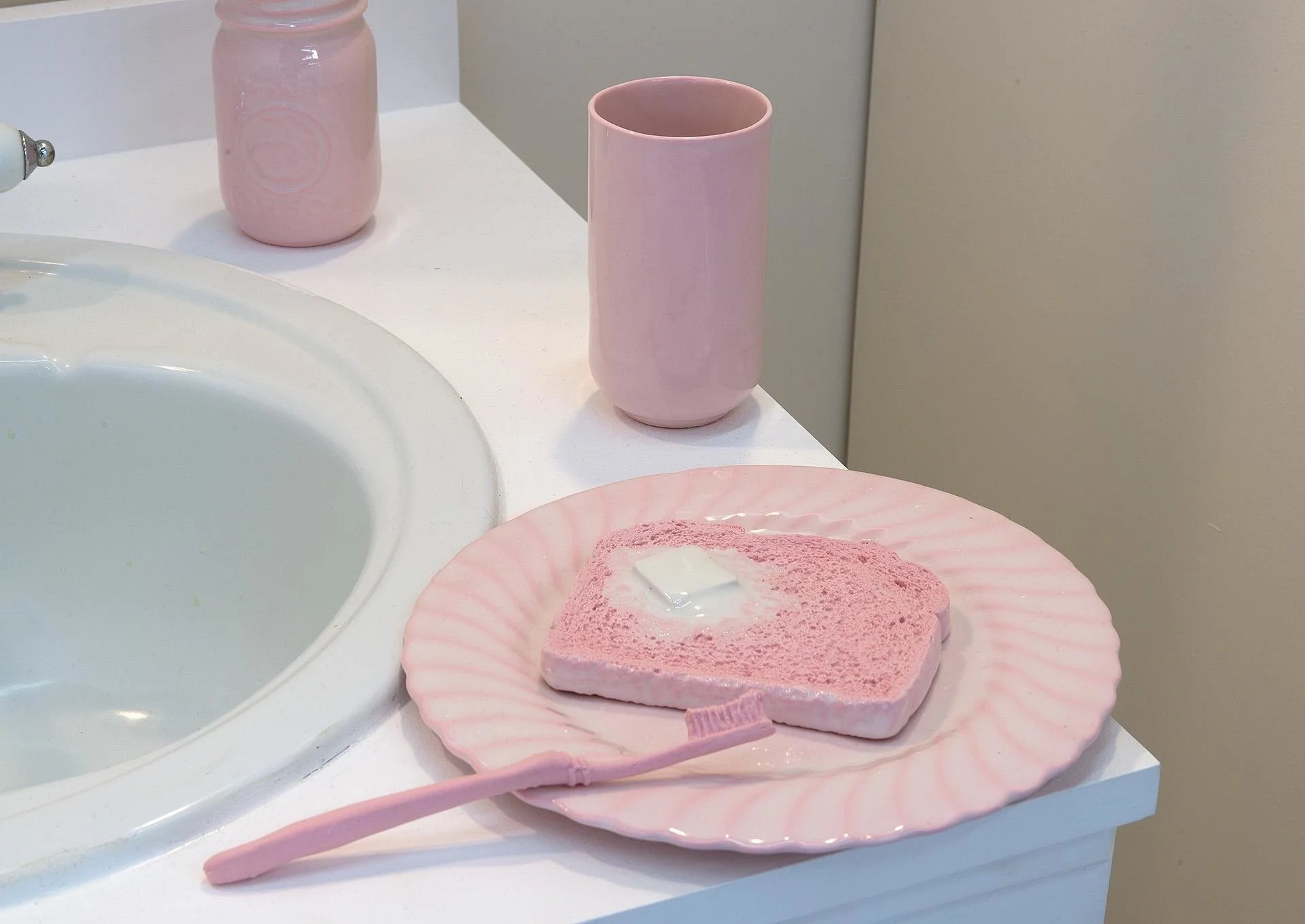 A pink porcelain piece of toast with a glazed pad of butter on a matching pink plate, with a matching ceramic toothbrush balancing on the edge. The plate is on the edge of a bathroom sink counter, with a matching pink cup and mason jar in frame.