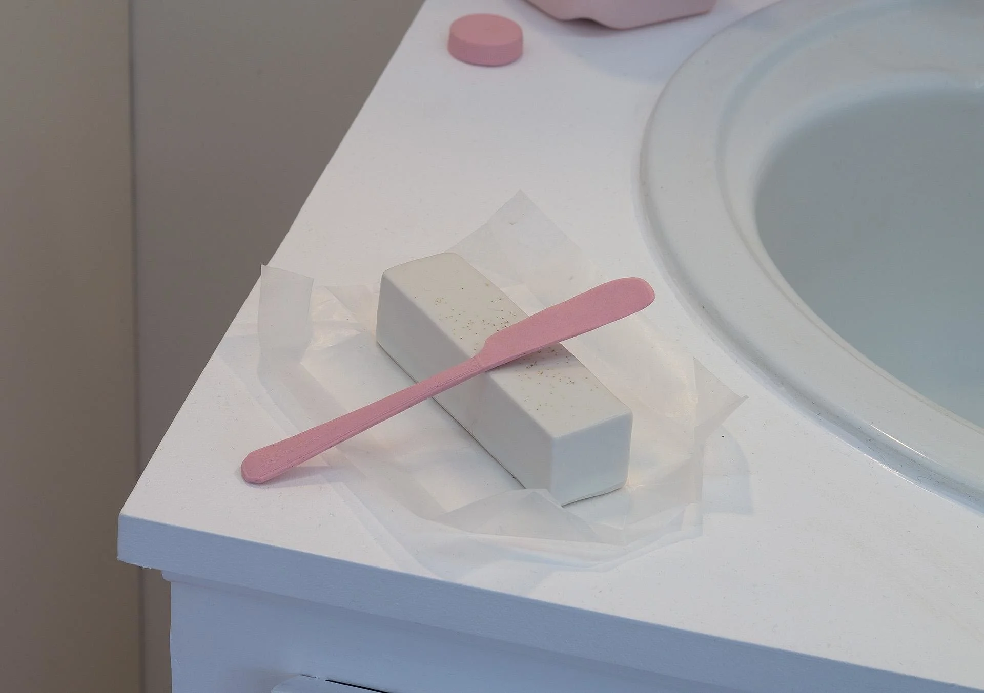A pink porcelain piece of toast with a glazed pad of butter on a matching pink plate, with a matching ceramic toothbrush balancing on the edge, both resting on the bathroom counter by the sink.