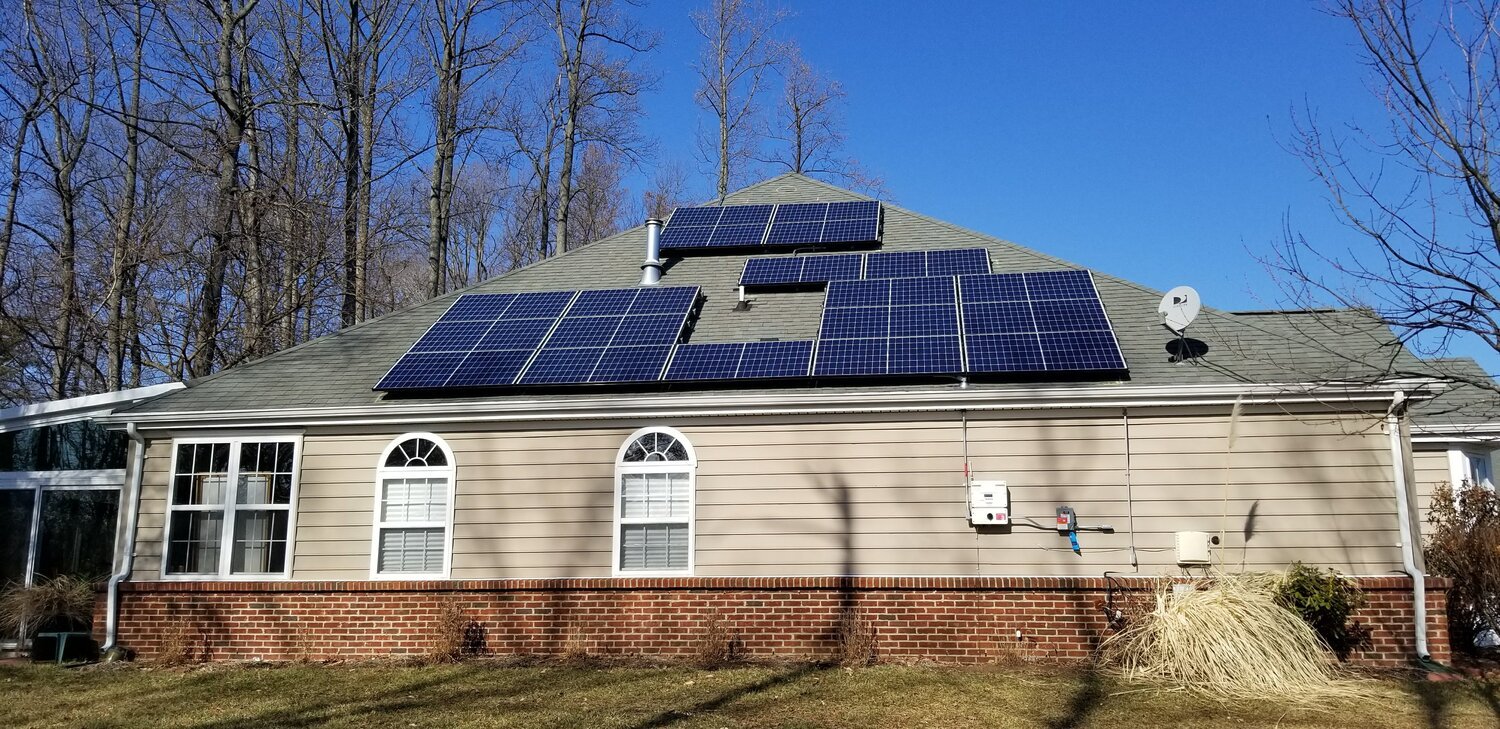 House with multiple solar panels on the roof, beige siding, brick foundation, two arched windows, satellite dish, and clear blue sky.