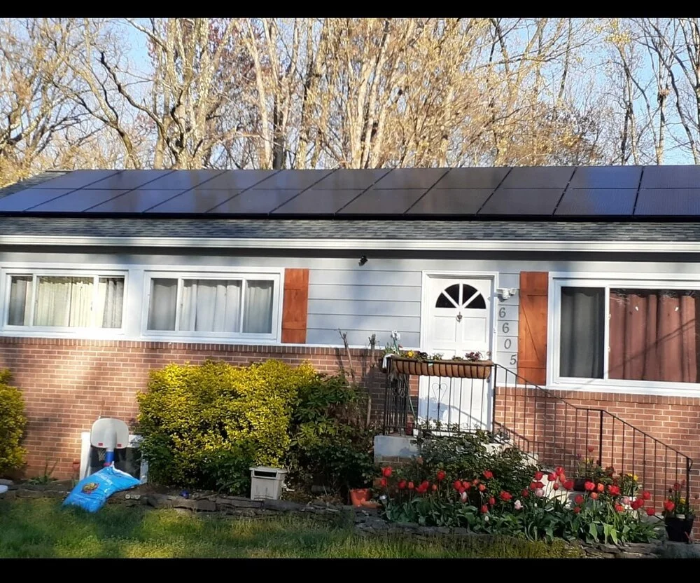 Front view of a suburban house apartment with a solar panel roof, brick and siding exterior, garden in the front yard with colorful tulips, bushes, and plants, a white door with house number 6605, stairs with a metal railing, and trees with bare bran