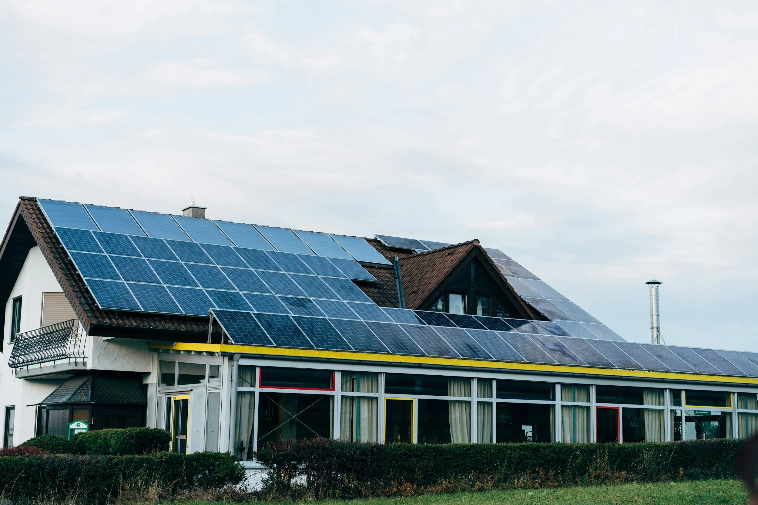A house with solar panels on its roof and a glass extension on the front, surrounded by a small hedge and a grassy yard.