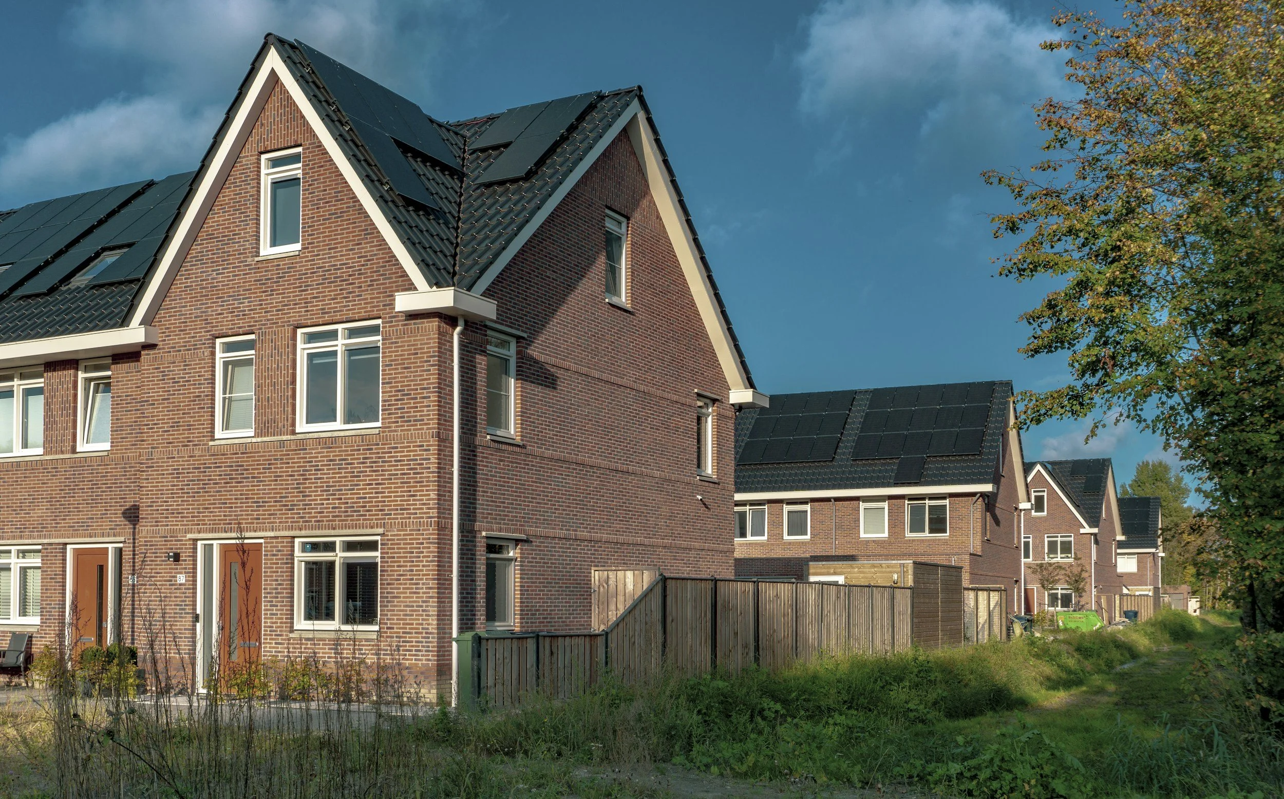 A row of modern brick houses with black roofs and solar panels in a suburban neighborhood, with greenery and a dirt path in the foreground.