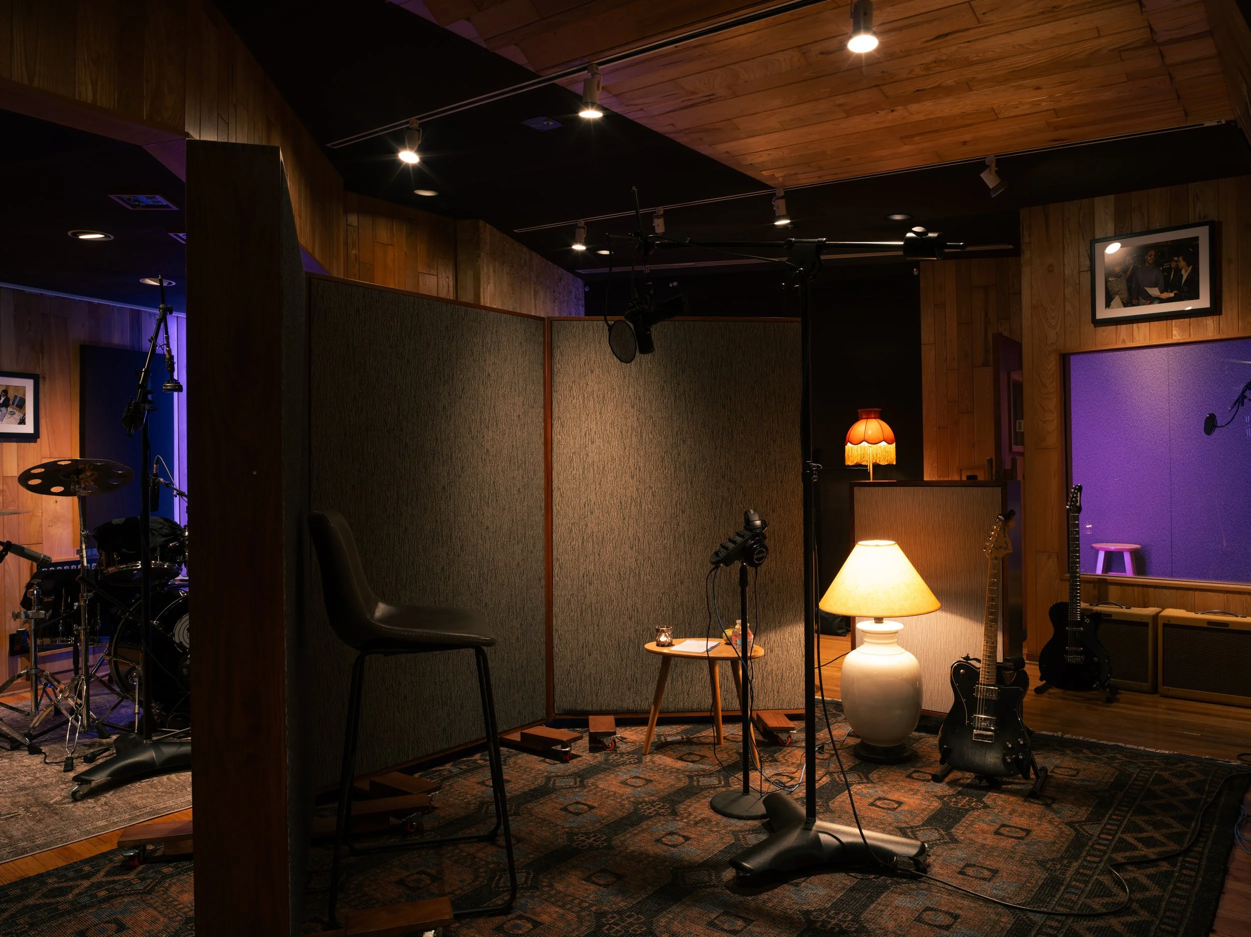 Recording studio with musical instruments, microphones, guitars, a drum set, and studio décor including lamps and framed photos on wooden-paneled walls.