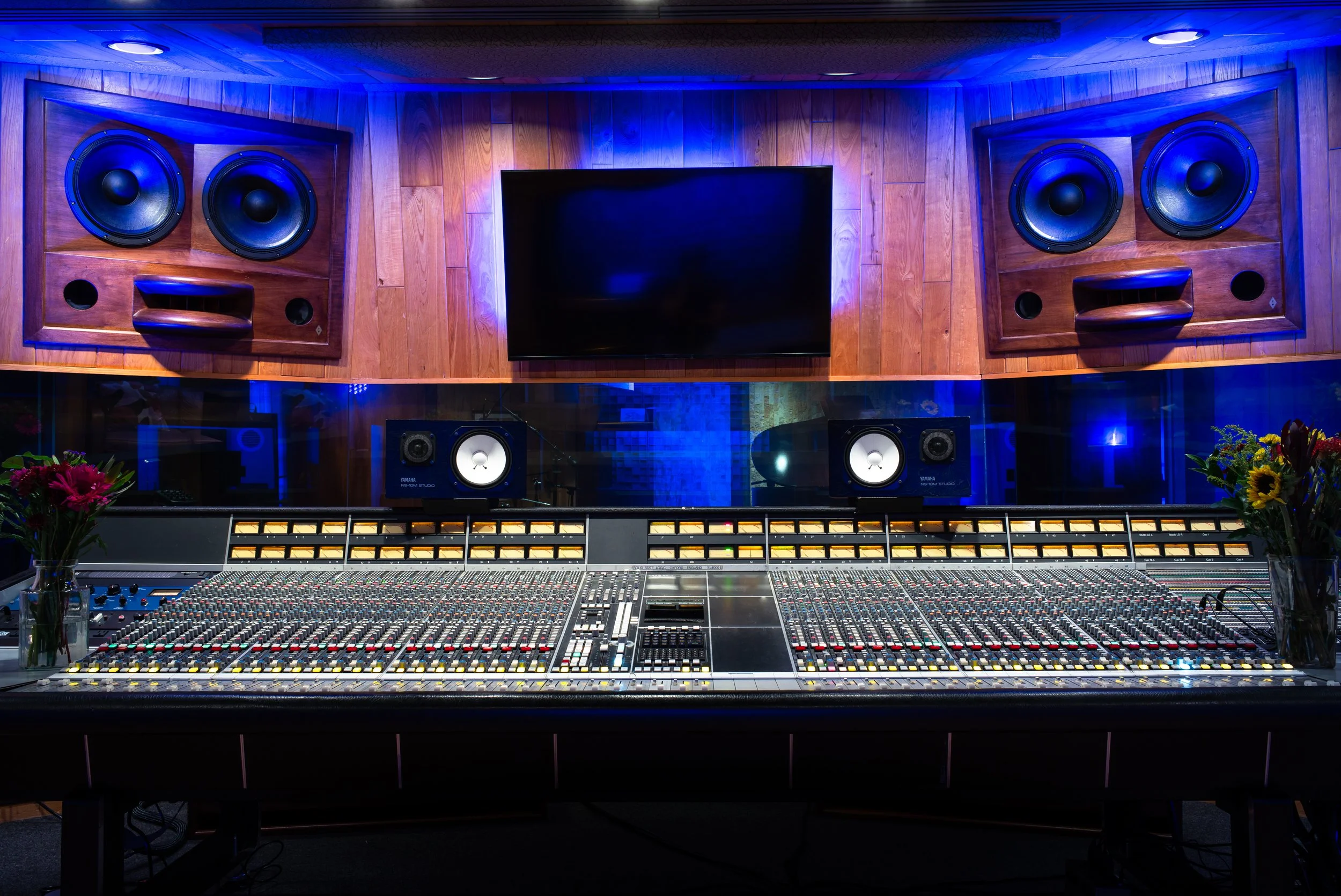Sound mixing console in a recording studio with acoustic panels, speakers, a large monitor, and floral decorations.