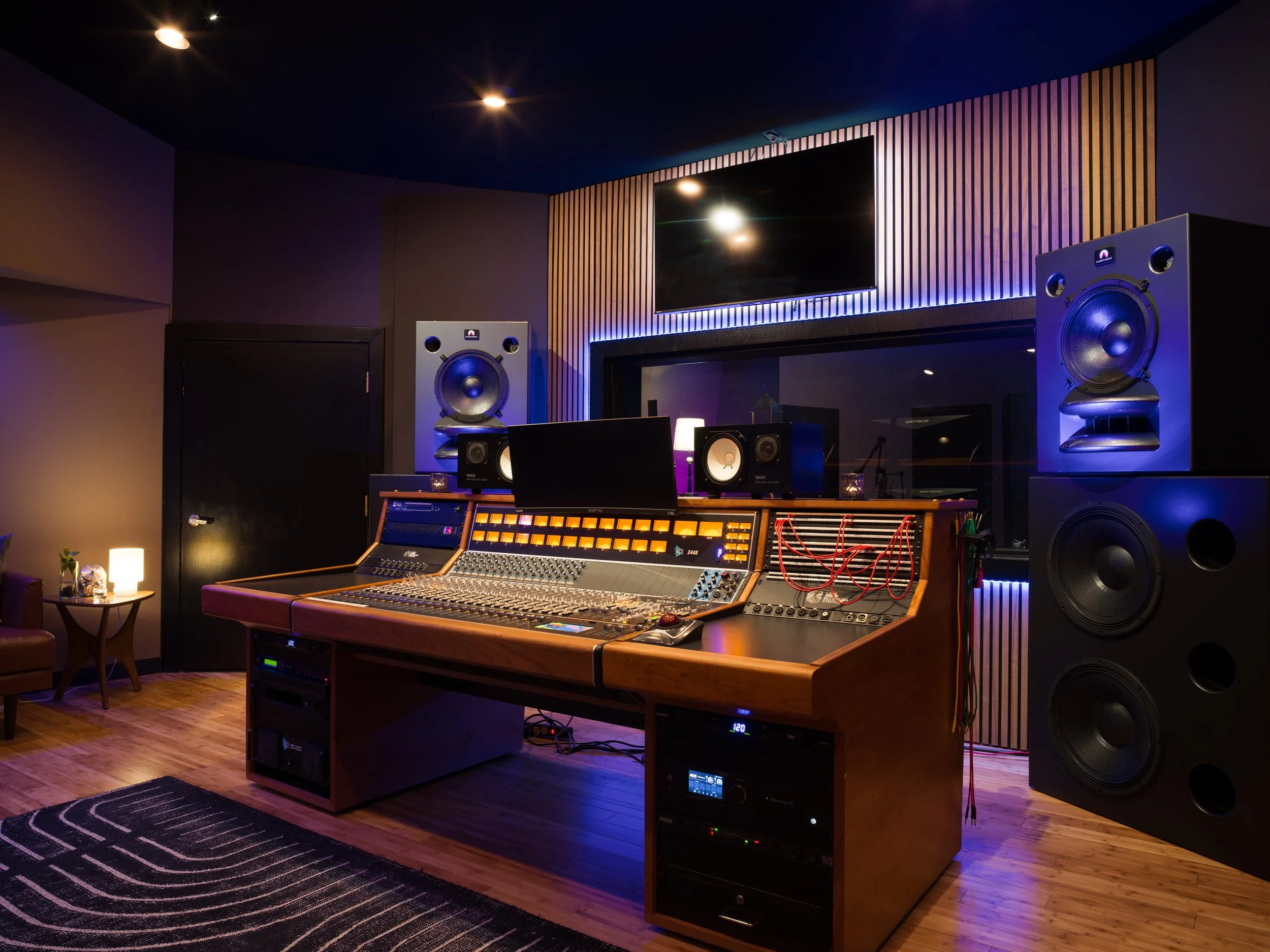 Recording studio with professional audio equipment, large speakers, mixing console, and soundproof window, illuminated with blue and warm lighting.