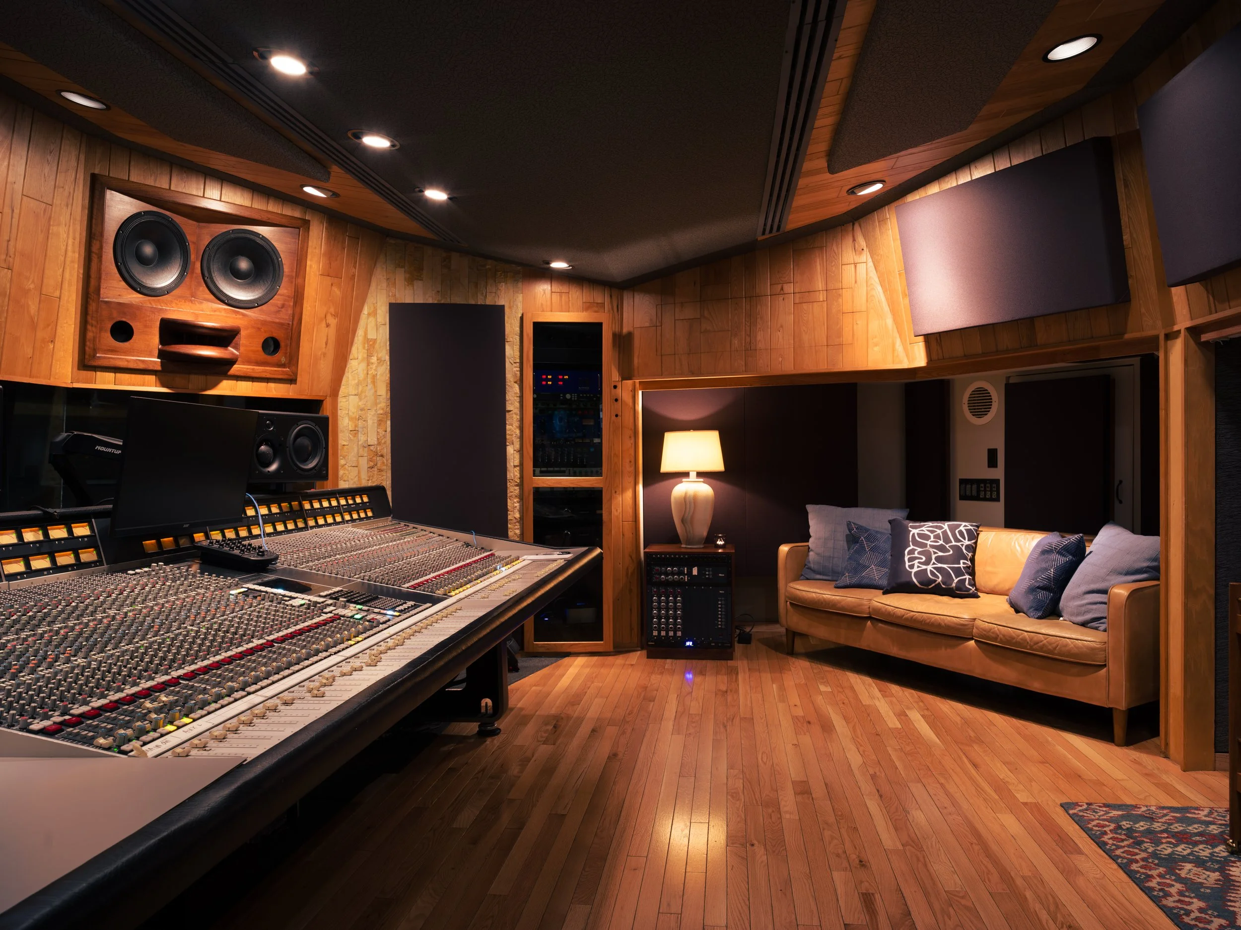 A professional recording studio with wooden walls, a large mixing console, studio monitors, soundproofing panels, a couch with pillows, a table lamp, and audio equipment.