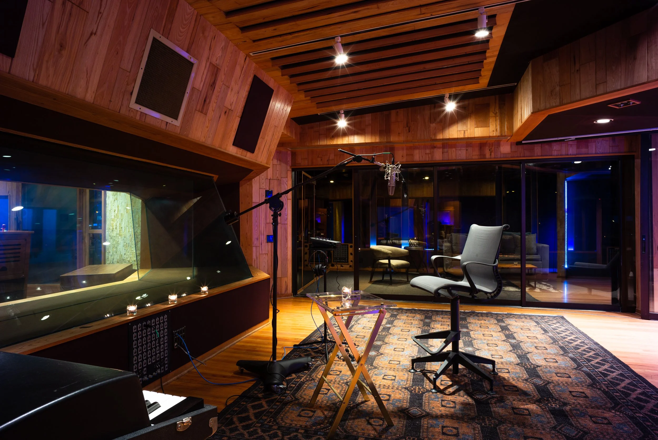 Recording studio with wooden walls, a large window into the control room with a chair, microphone on a stand, and recording equipment.