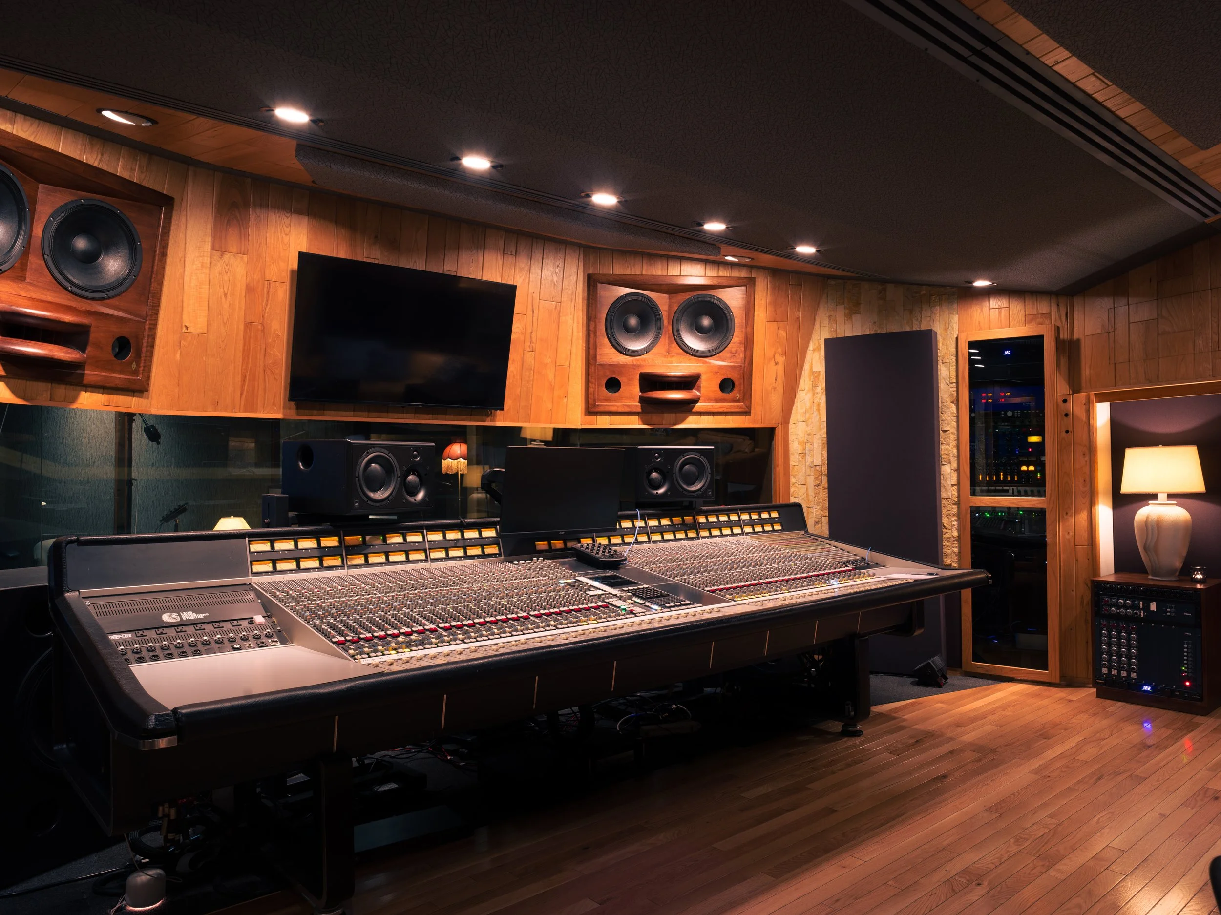 Recording studio with a large mixing console, multiple speakers, a television screen, sound equipment, wooden walls, and dim lighting.