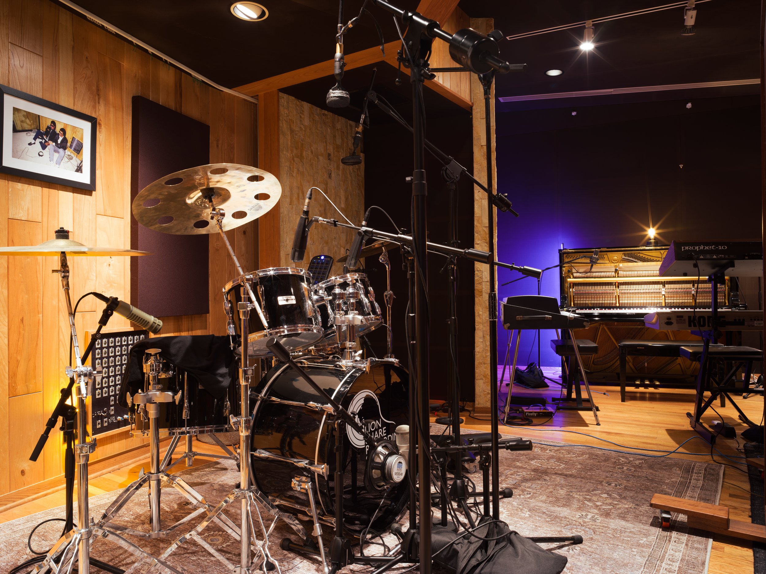 Music rehearsal studio with drums, keyboard, microphones, and music equipment set up, illuminated with stage lighting.