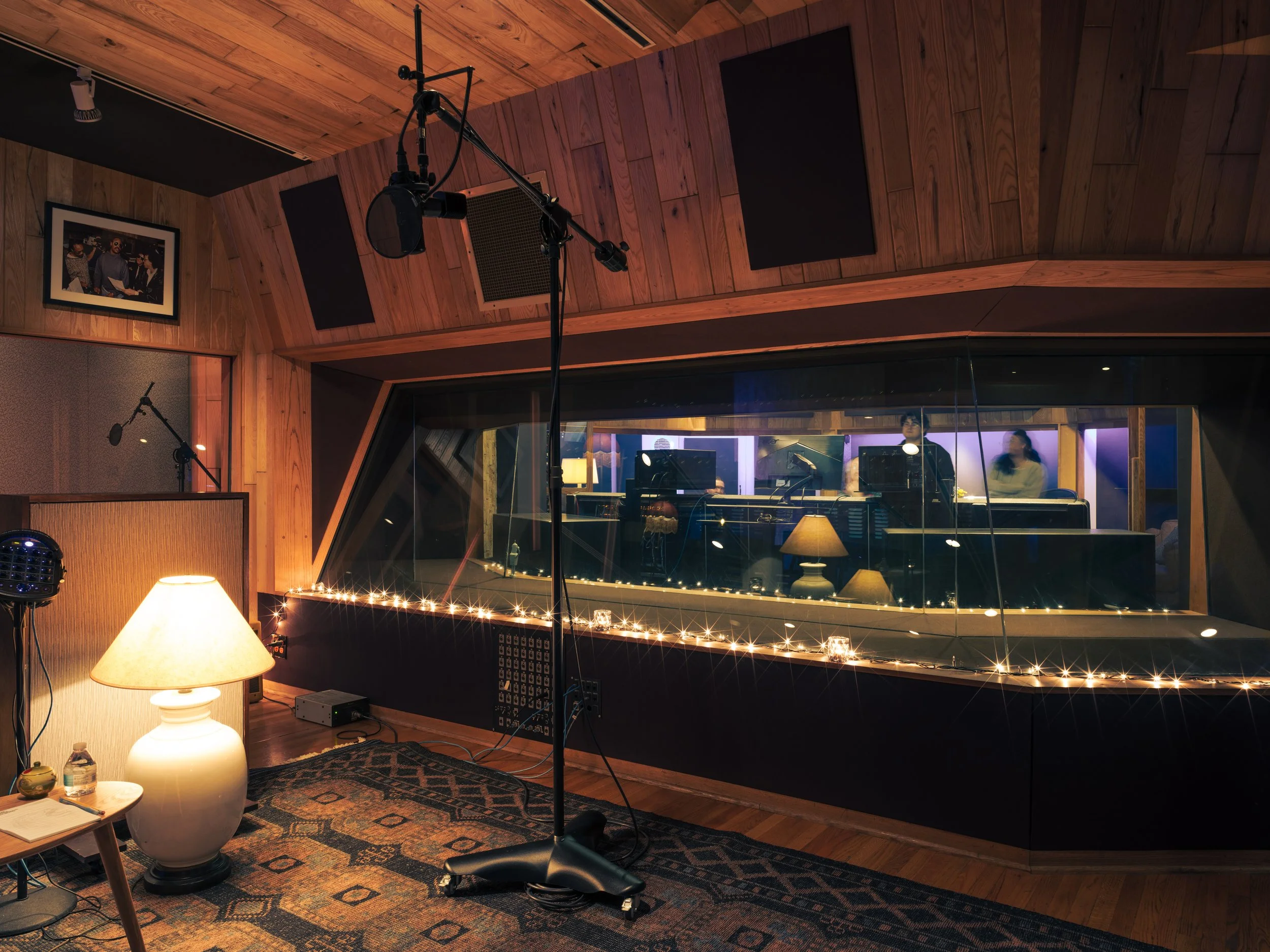 Recording studio with a large window overlooking the control room where two people are working at the mixing console. The room has wooden walls and ceiling, a lamp on a side table, and string lights along the window ledge.