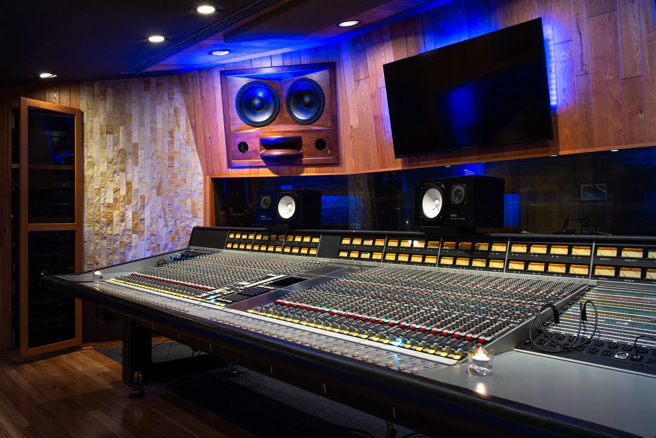 A professional recording studio with a large audio mixing console, multiple screens, and studio monitors, with wood-paneled walls and blue accent lighting.