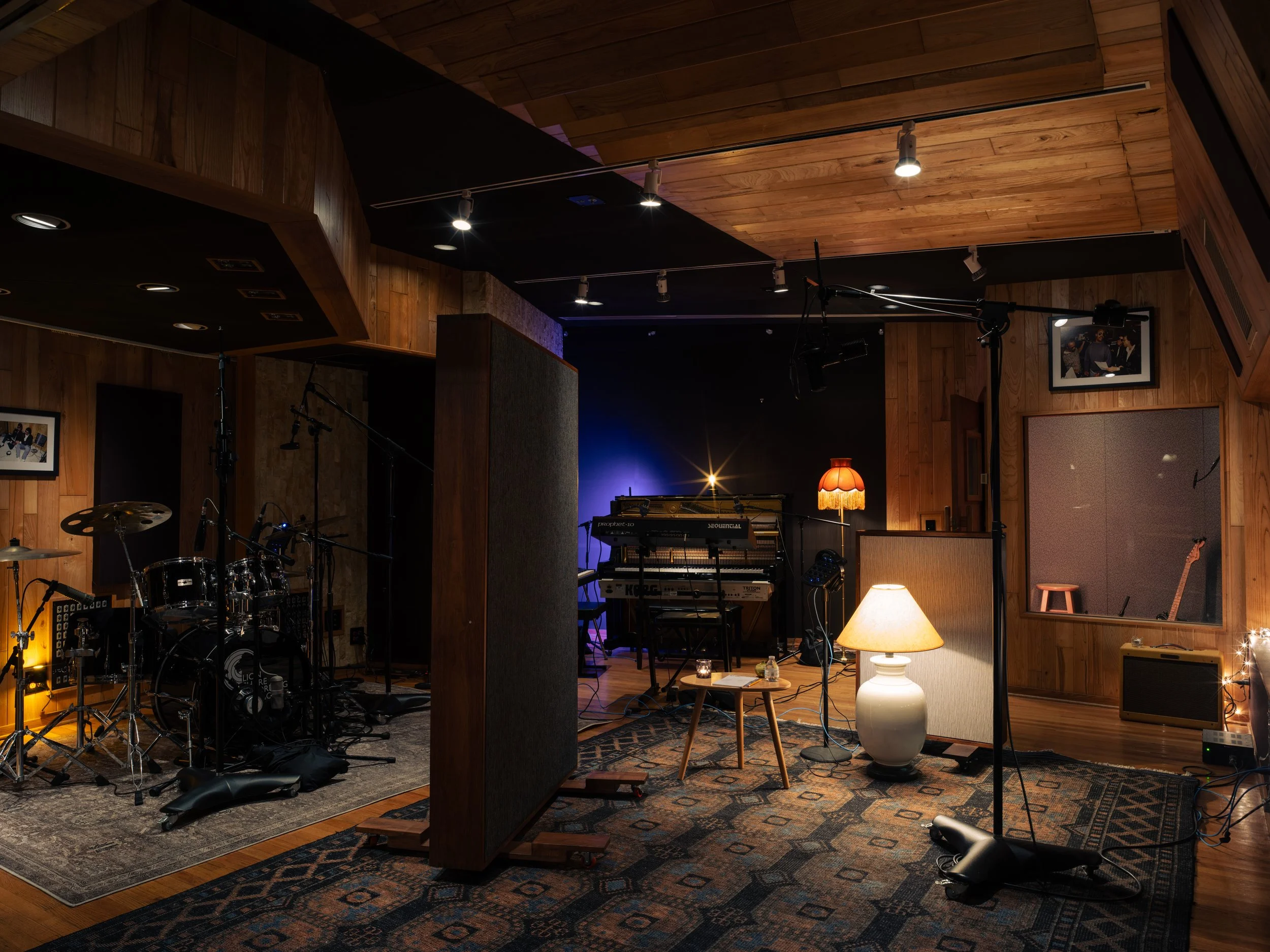 Music studio with drum set, keyboards, microphones, acoustic panels, lamps, and musical instruments on wooden walls and carpeted floor.