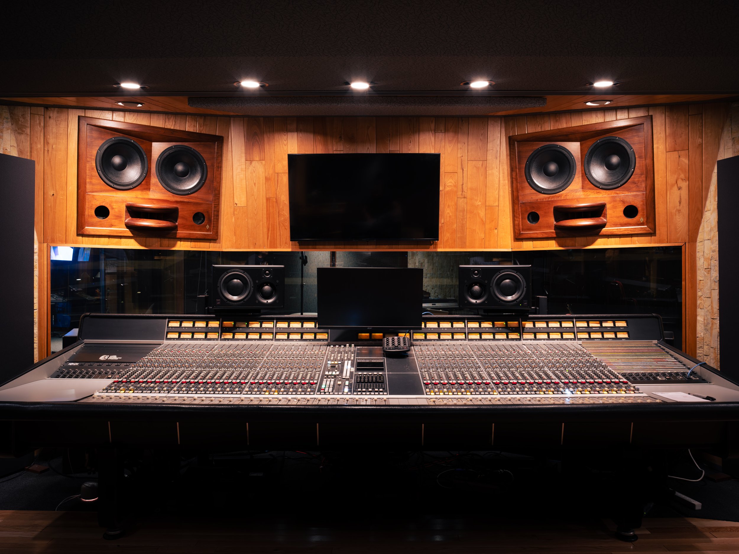 Recording studio with a large audio mixing console, surrounded by professional speakers and a wooden-paneled wall with a flat-screen monitor.