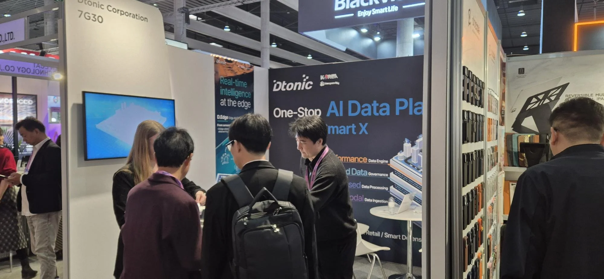 At a technology trade show, a booth displays banners for Dtonic's AI Data Platform Smart X, with people engaging in conversations. The booth features a wall of various tech devices or accessories, and a monitor screen.
