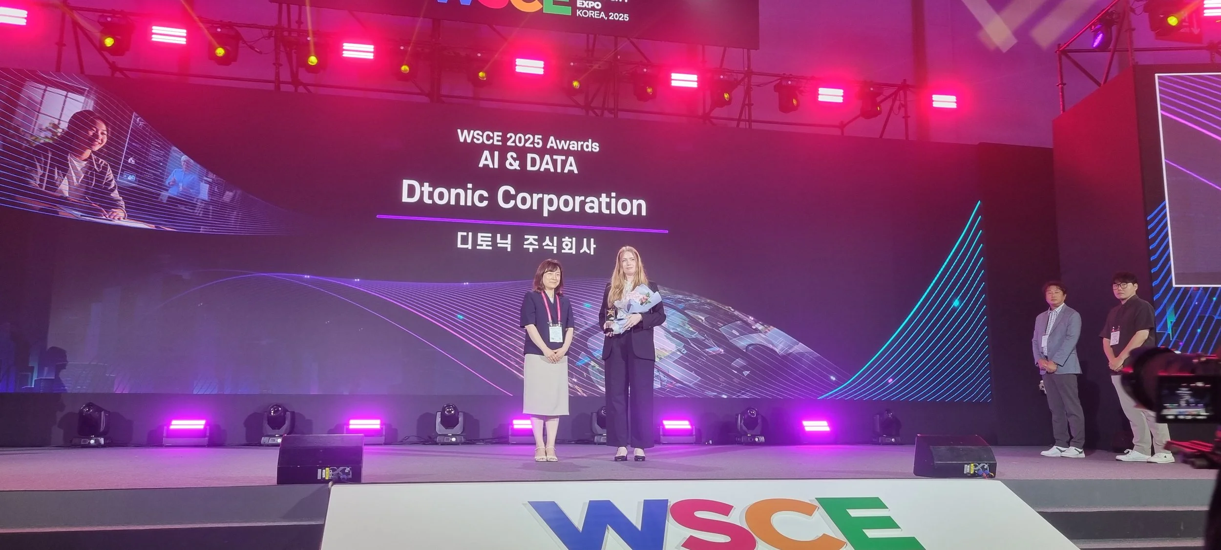 At the WSCE 2025 Awards event, a woman is receiving an award from another woman on stage. The backdrop displays the event details: 'WSCE 2025 Awards AI & DATA Dtonic Corporation' in both English and Korean. Pink and purple stage lighting illuminates 