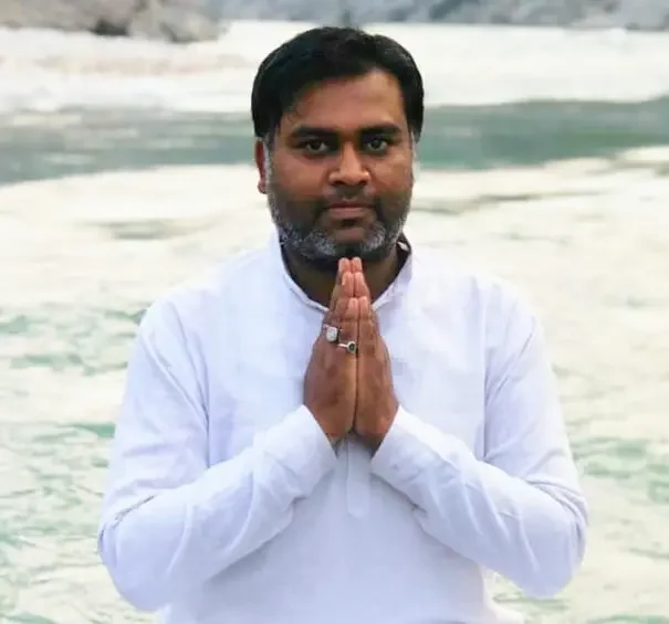 A man with dark hair and beard, wearing a white shirt, with hands folded in a traditional Indian greeting, standing near water with waves in the background.