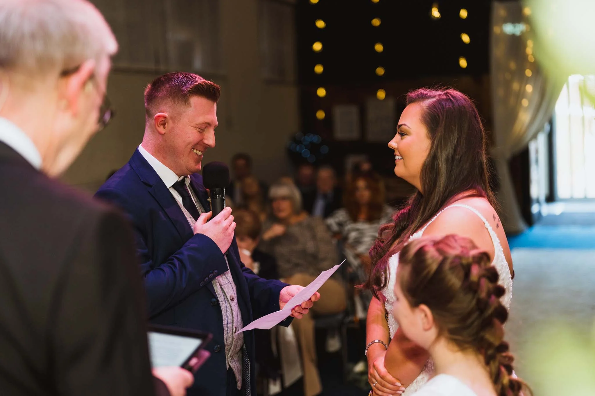 The groom makes a speech in front of the bride, with guests in the background and the minister in the foreground.