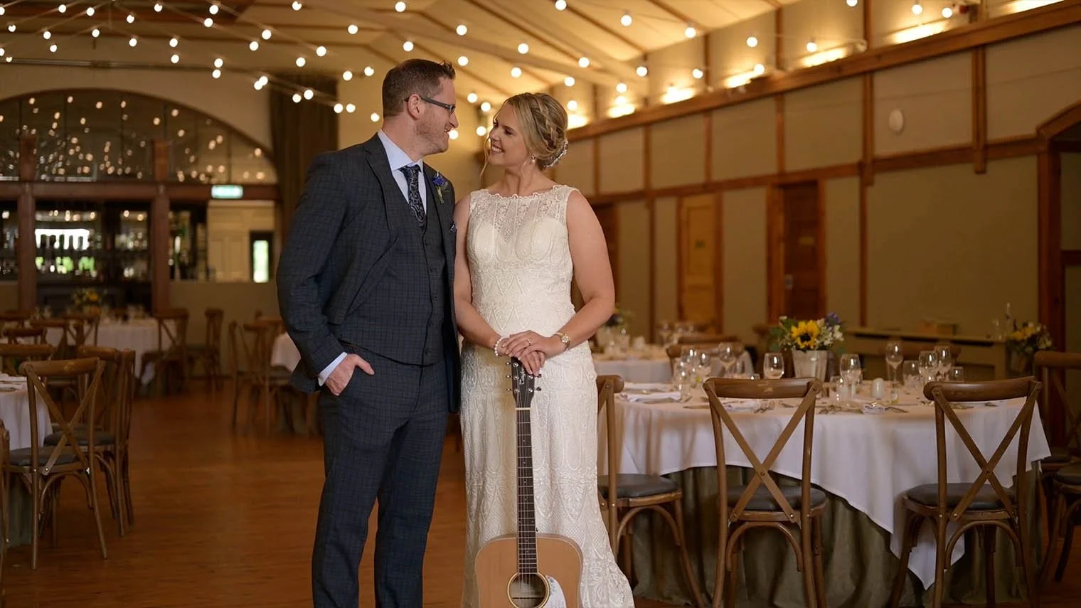The bride, holding a guitar, looks at the groom in the reception room.