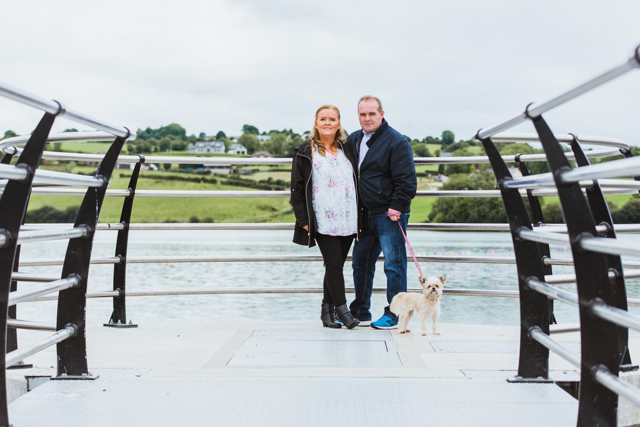 An older couple and a dog with the water and countryside in the background.