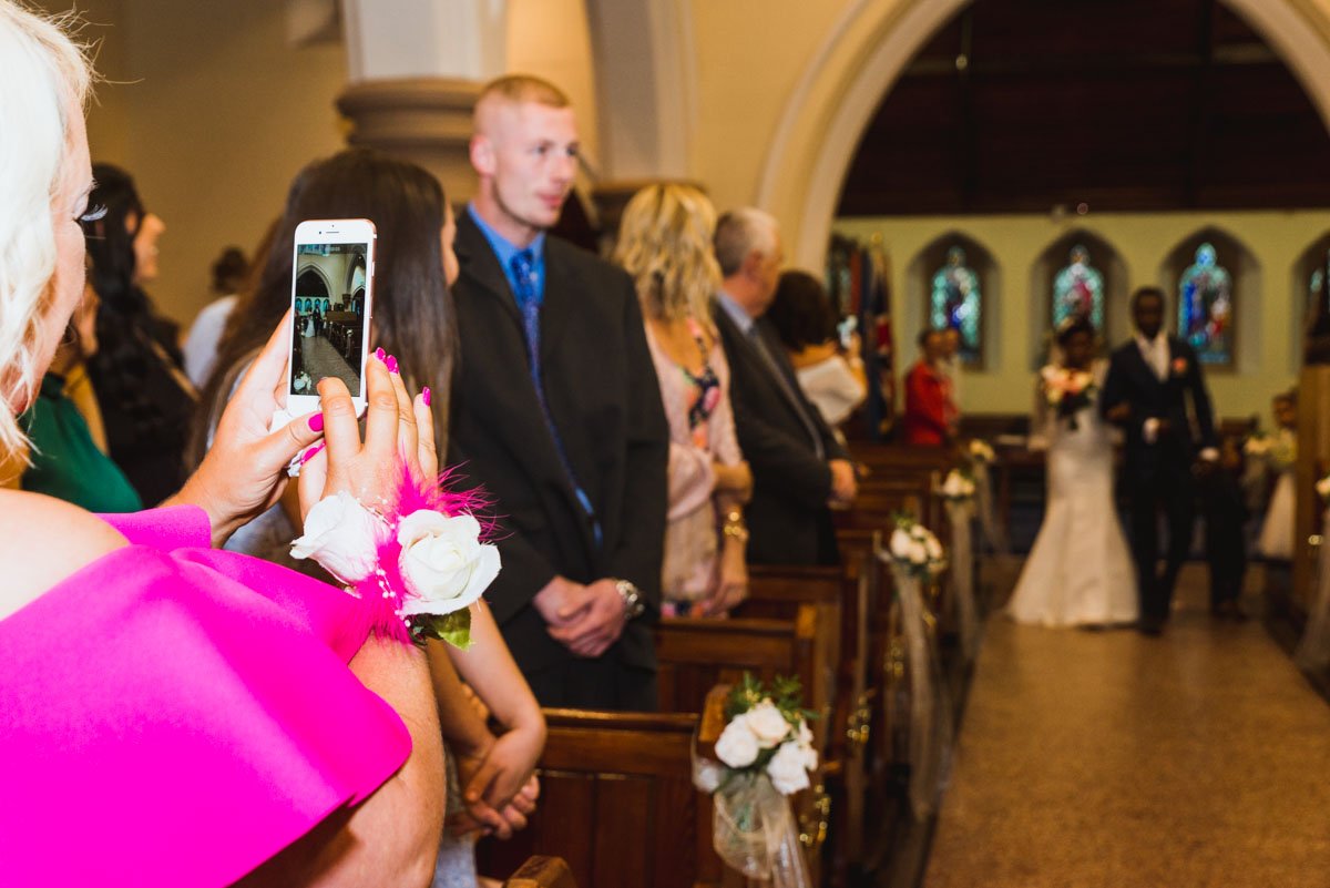 Bride with brother walk down the aisle as someone in the foreground is taking a photo on their phone.