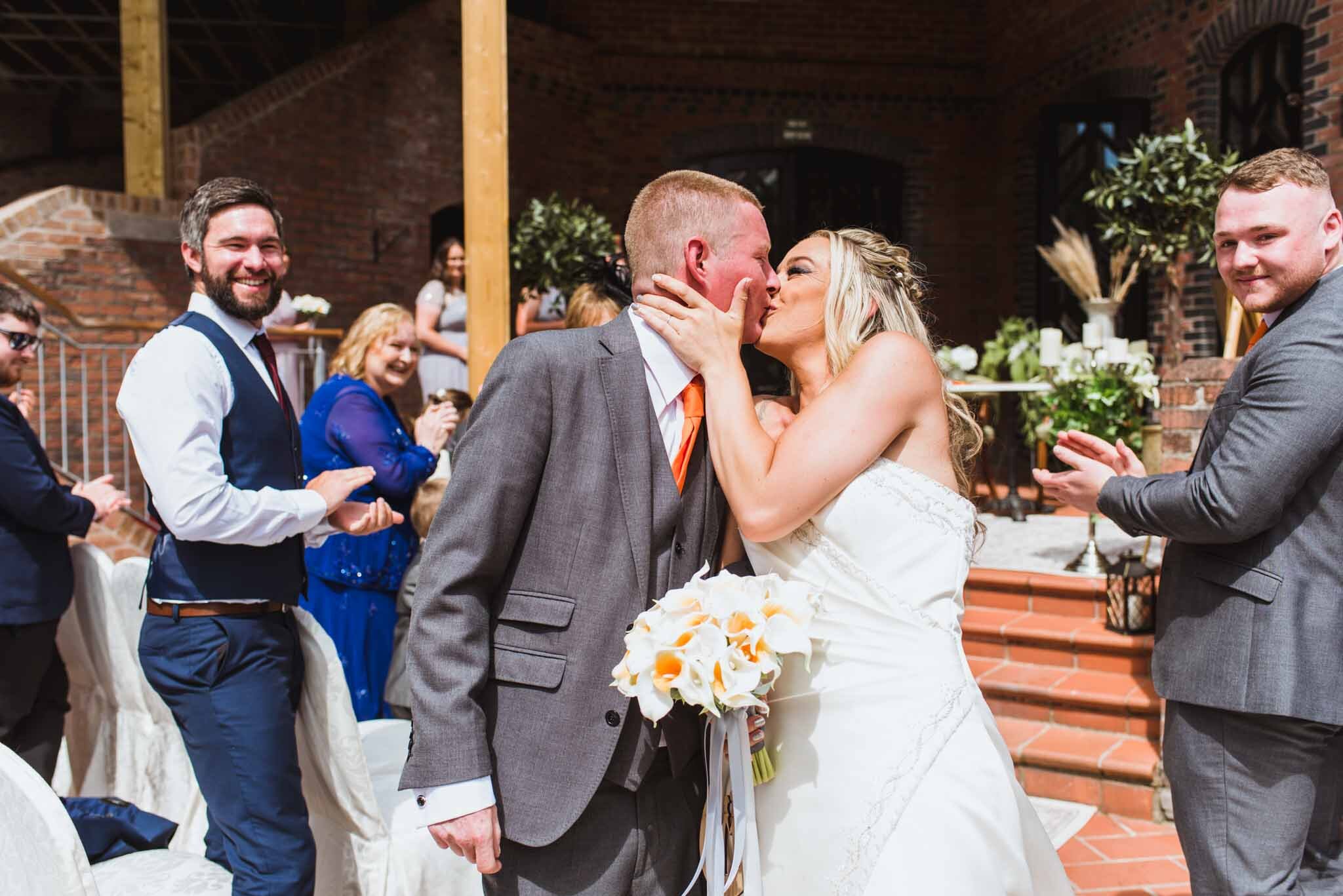 The bride and groom kiss at the end of the aisle with guests behind cheering and happy after the ceremony. 
