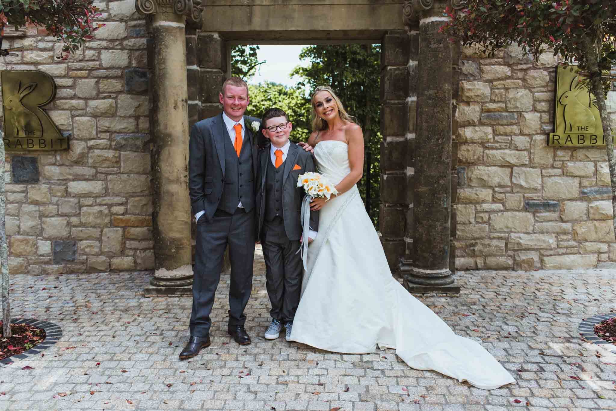  The bride, groom and their son are in front of a stone arch at the back of the hotel. 