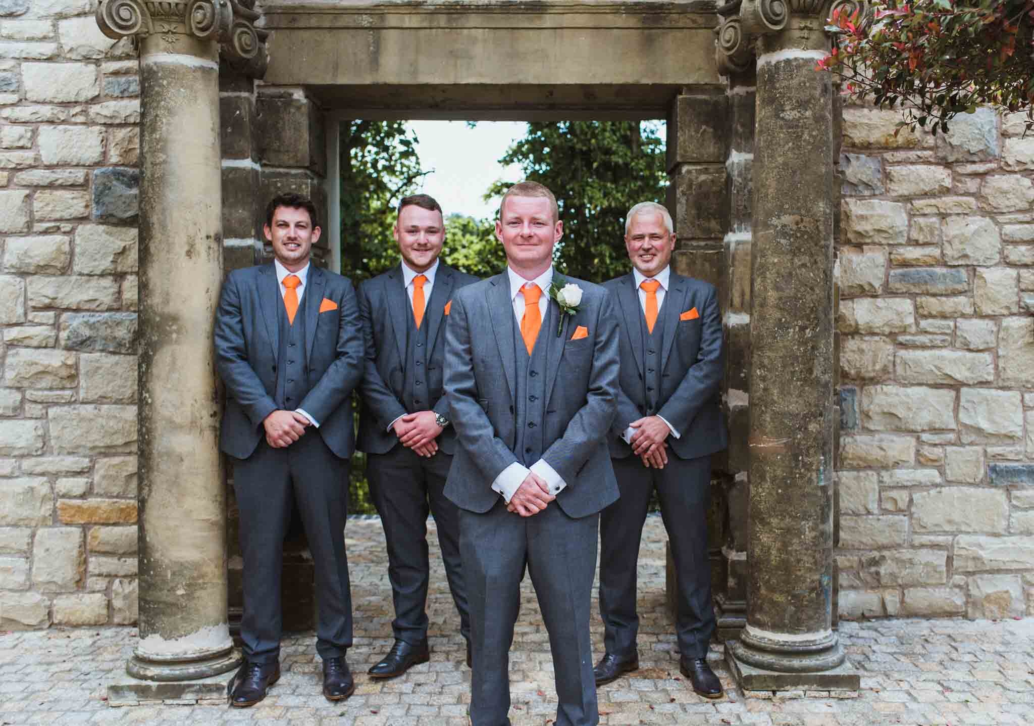  Groomsmen with groom slightly in front holding hands in front of arches and pillars in hotel gardens. 
