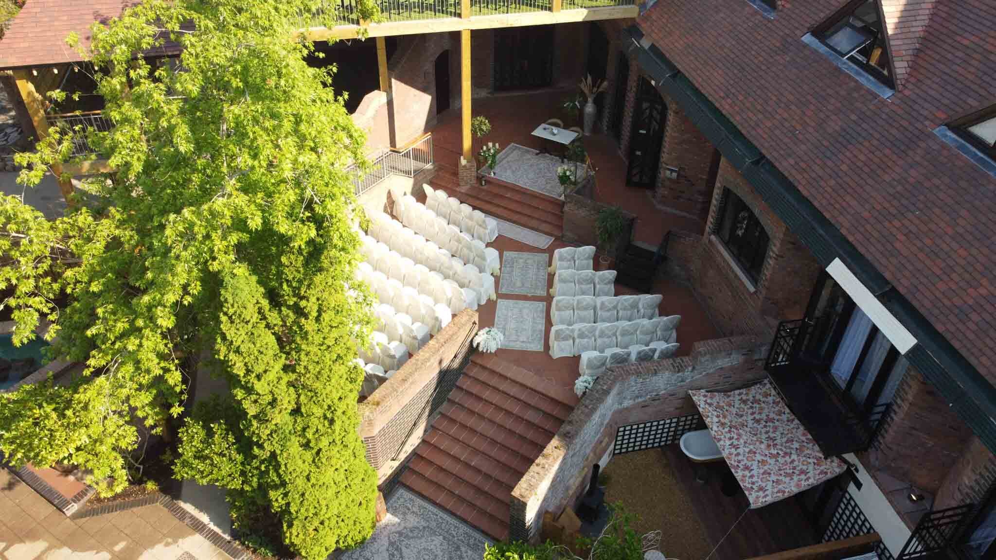  Drone bird's eye view looing down the ceremony at the back with empty chairs waiting for guests to arrive with trees to the left and steps to the garden. 