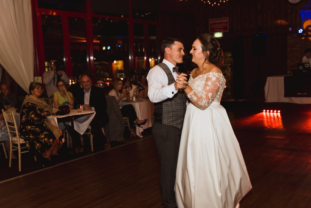  Slow first dance with bride and groom holding each other at their evening reception in the Riverdale Barn in Aghagallon as is music playing.  Guests watch them dance. 