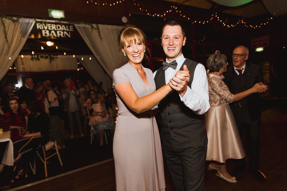  The maid of honour and best man dancing holding hands looking at the camera in a dark environment with other guests dancing behind. 