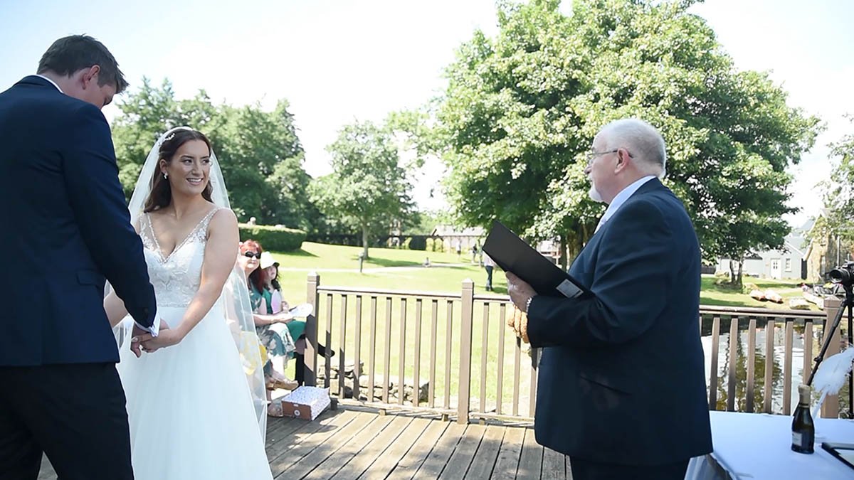  Outside in the sun humanist celebrant John Foley conducts a ceremony holding a book with the bride and groom to the left and trees off in the distance. 