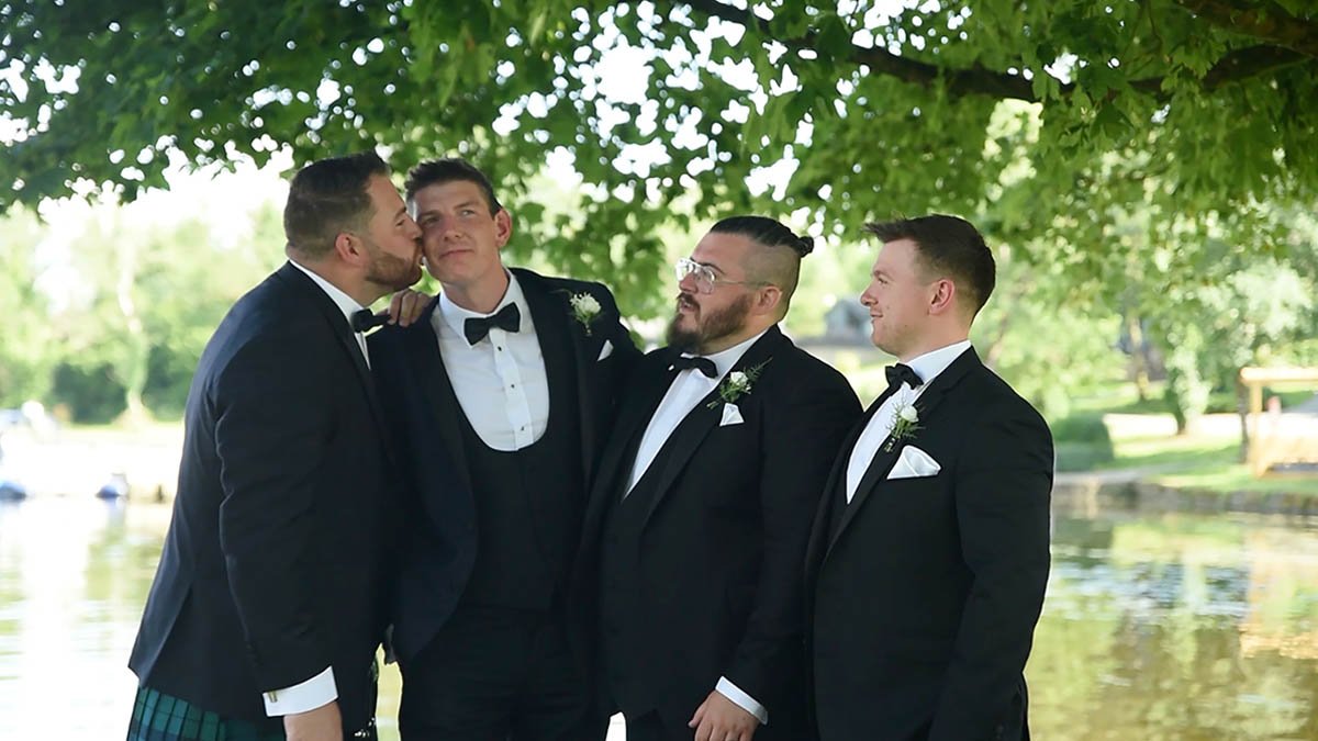  Groom with groomsmen having fun chatting under trees just outside venue on a summers day wedding. 