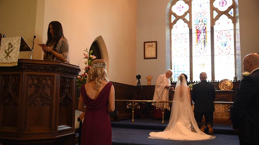  The ceremony in the church as female reads as the bride and groom kneel and the priest is at the front of the church. 