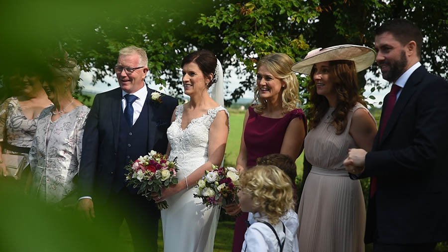  Family outside in the garden beside the church as the people stand side by side with the bride holding a bouquet. 