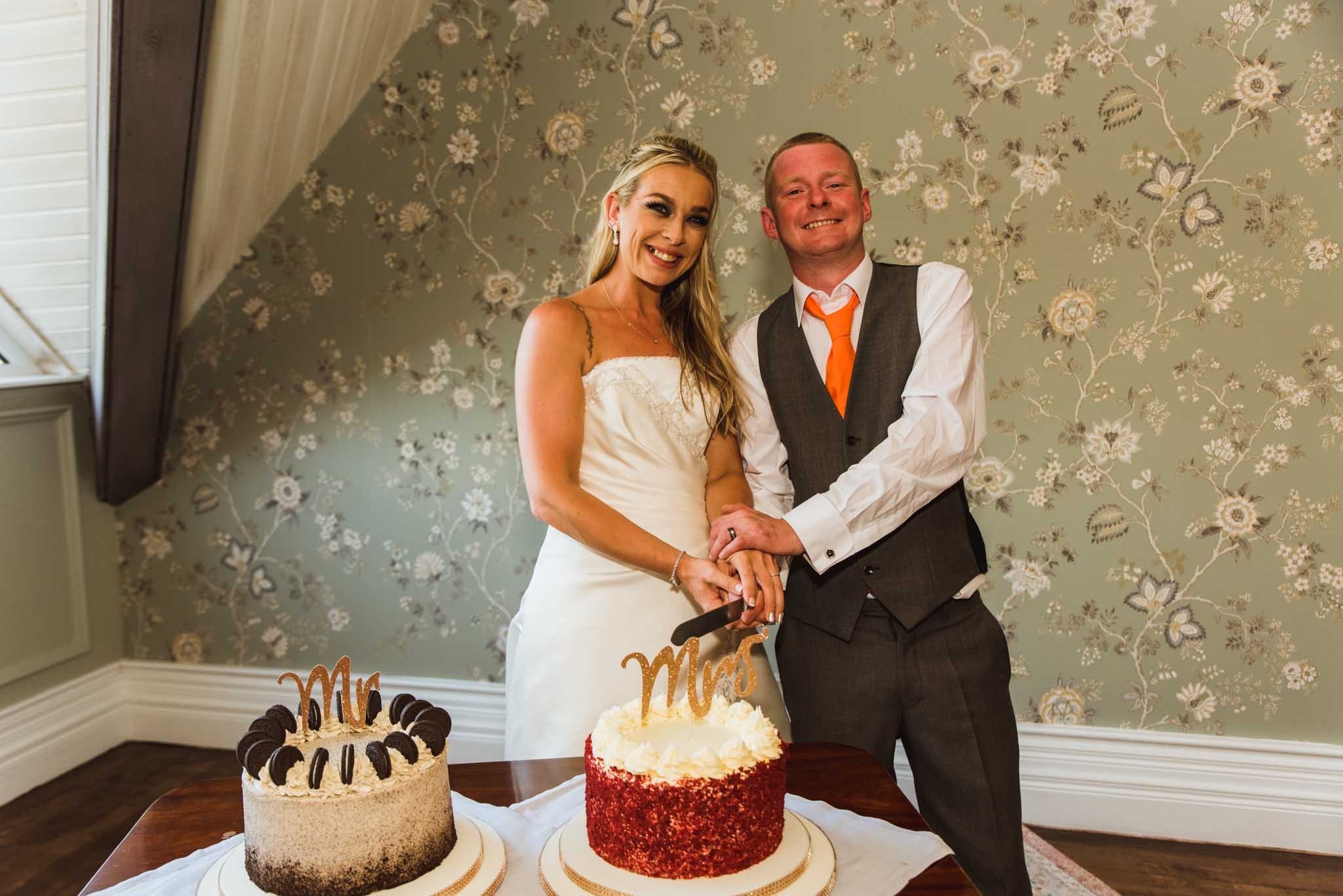 The bride and groom cut the wedding cake in the corner of the reception room with both hands on knive looking at the camera. 