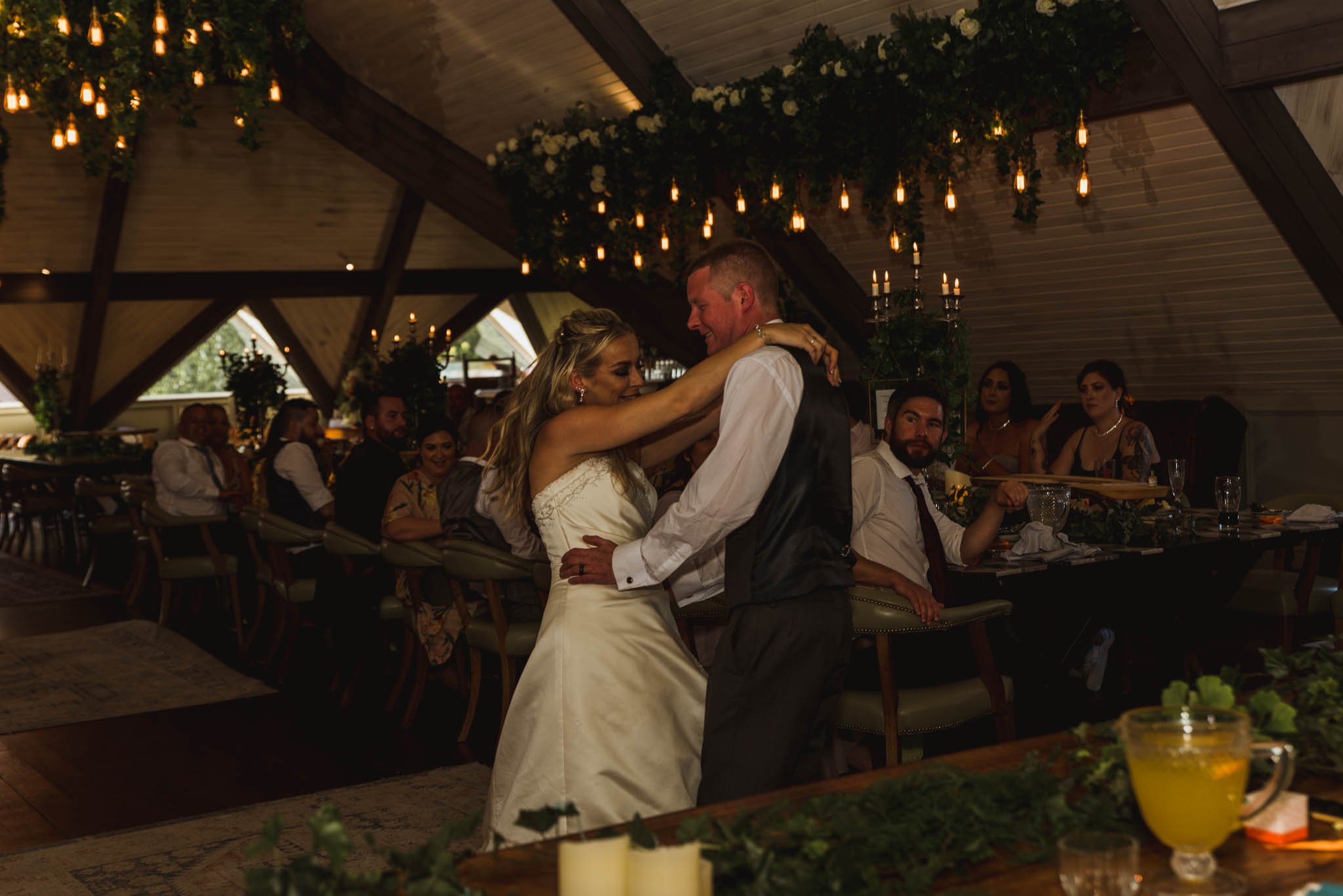  Brdies arms around her husband's shoulders while his hands are on his wife's hips as they slow dance in the dimly lit reception room upstairs with candles and fairy lights giving the room some atmosphere. 