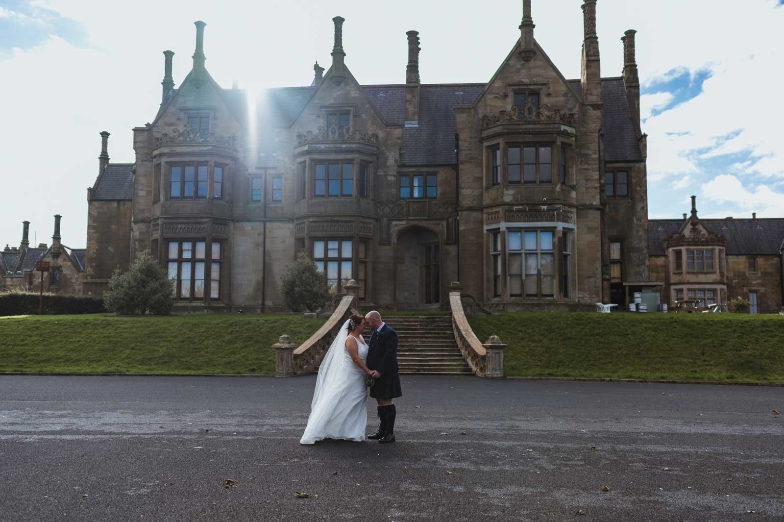  Wide photo of the bride and groom kissing in front of Brownlow Castle with the sun beaming with rays of light behind the building. 