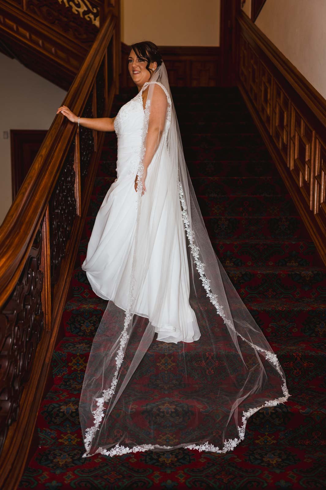 Full bride’s wedding dress as she is standing at an angle inside stairs with one hand on the banister. 