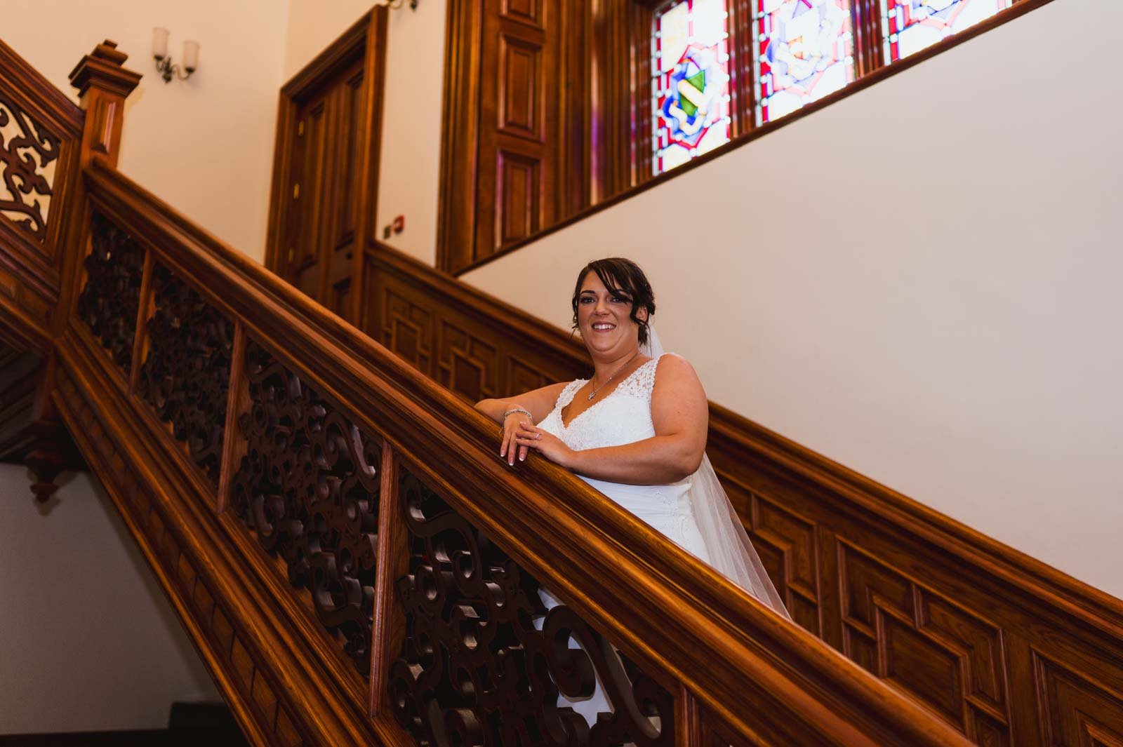  Bride leaning against wooden bannister on stairs within building showing some windows. 