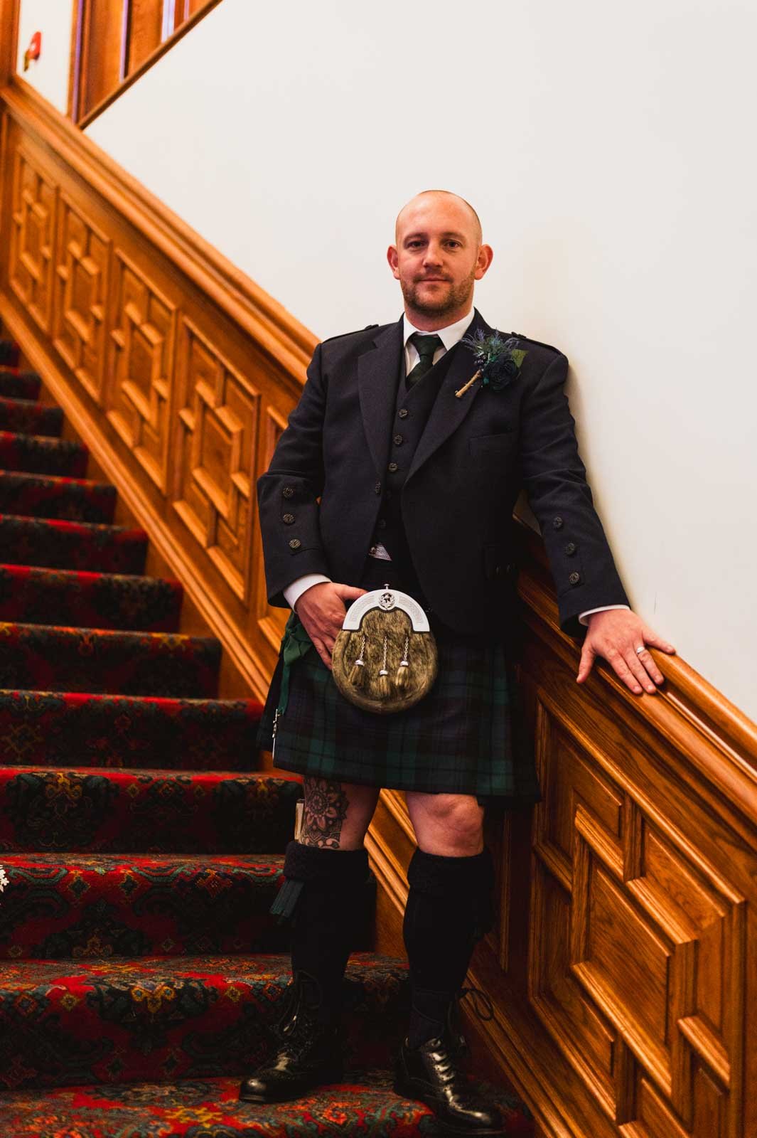  Grooms showing off Scottish wedding attire standing on the stairs with one hand on the railing. 