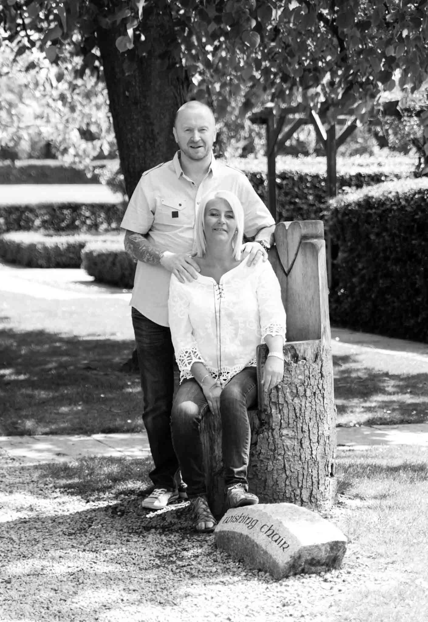  A female sits on a wooden carved heart chair as a male stands behind her with his hands on her shoulders. 