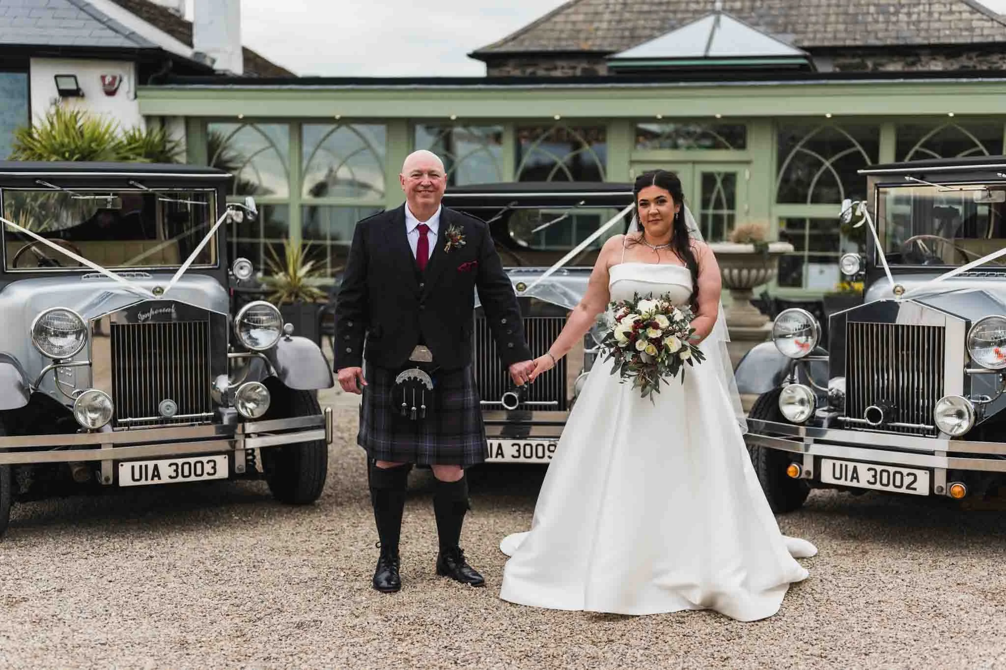 The bride is holding a bouquet, and the groom is holding hands, smiling in front of the limos outside the venue.