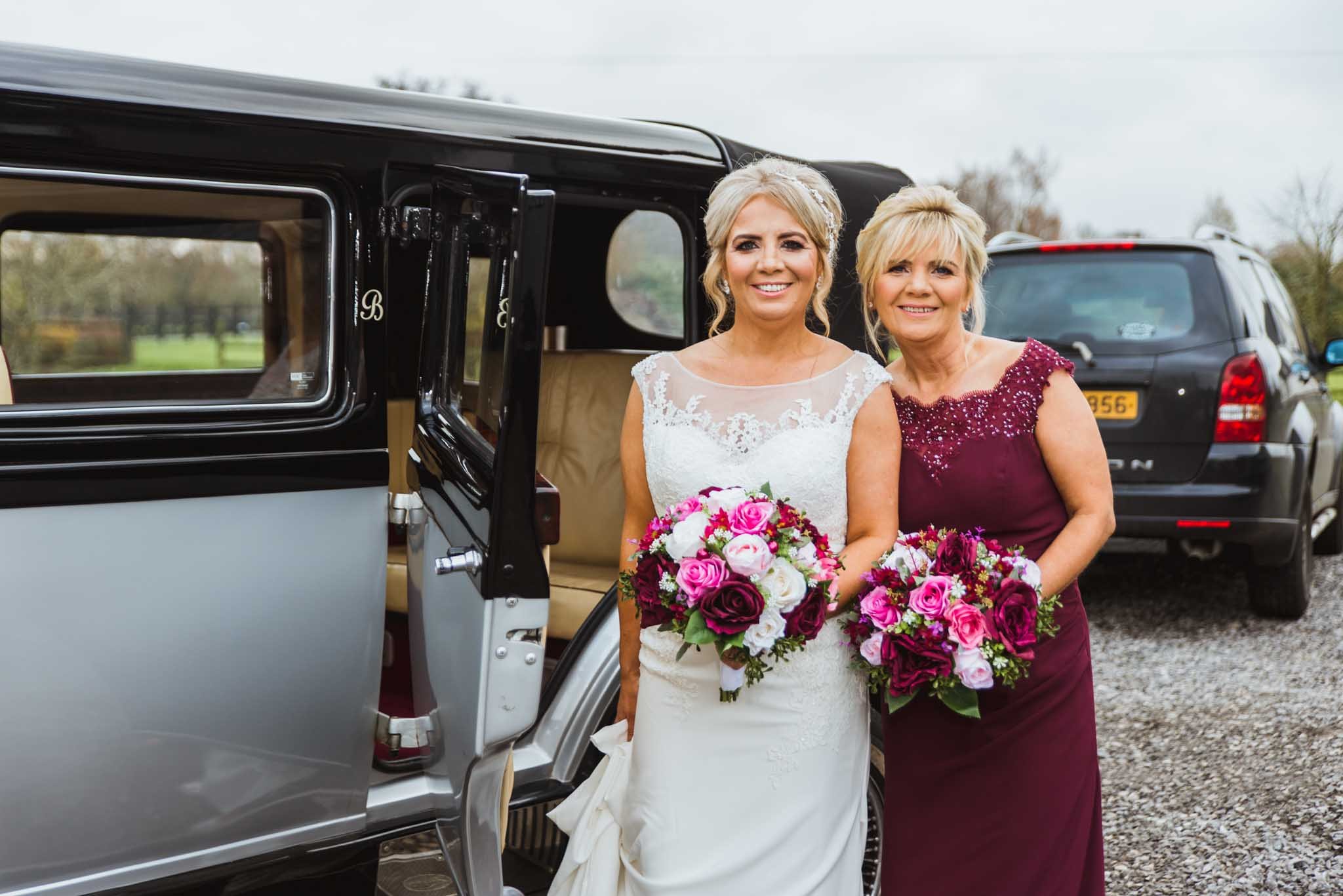 The bride and maid of honour stand beside the car with their bouquets.