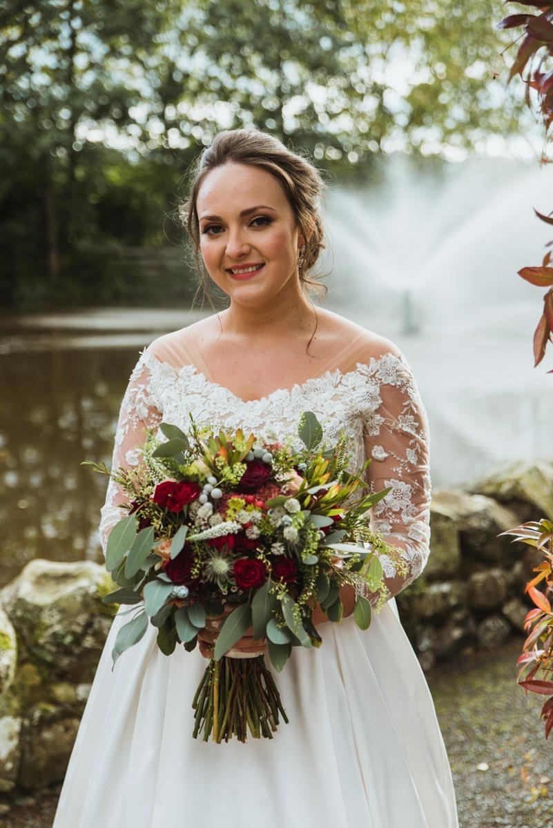 Bride holding a bouquet, looking at the video camera in front of a small stone wall with water spraying into the pond behind.