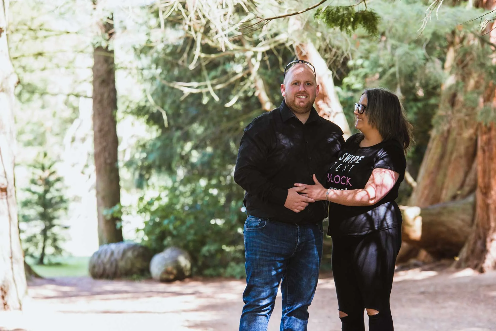 The female embraces the male while he looks at the camera, with plenty of trees around the older couple.