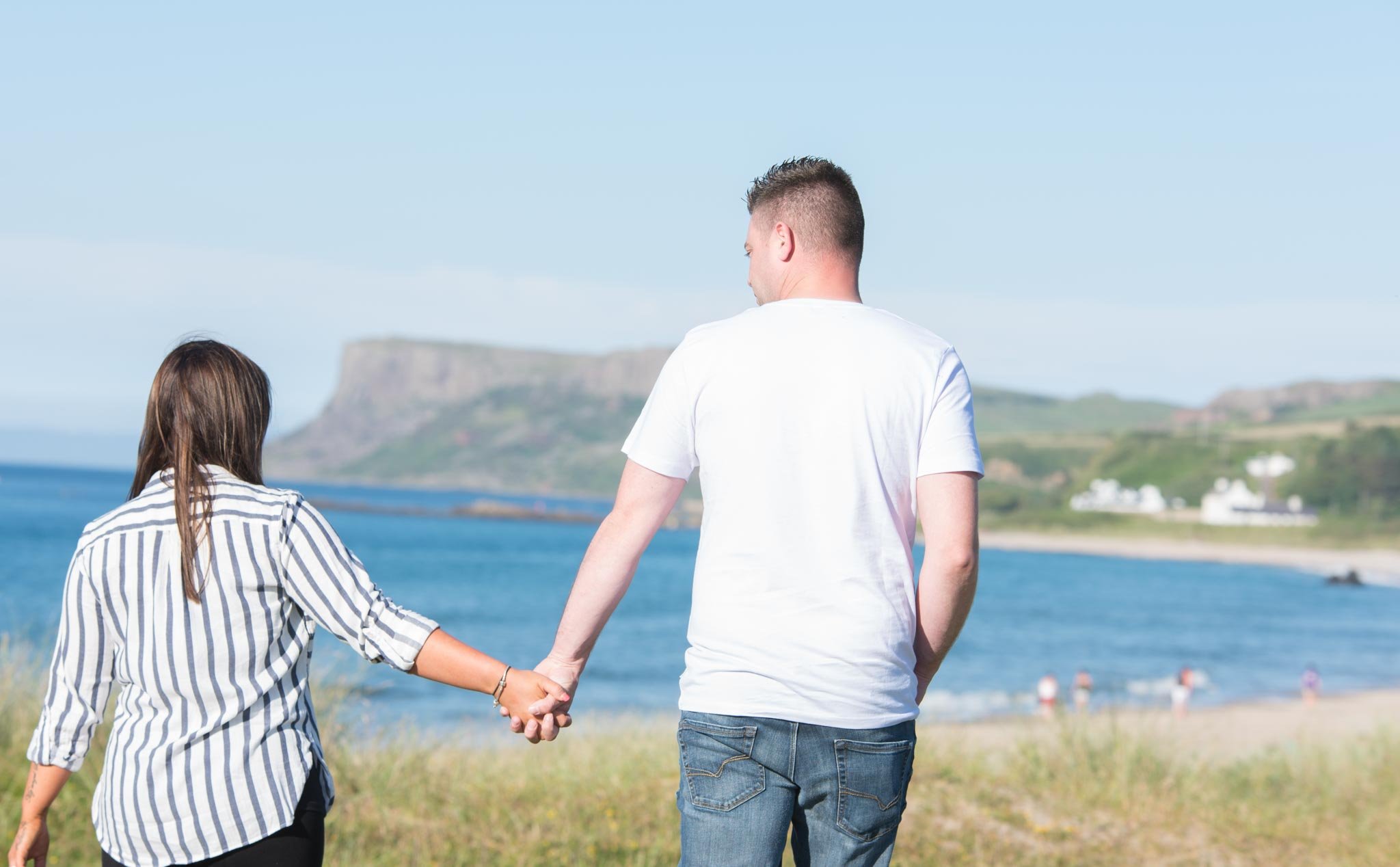 A couple holding hands is photographed from back with the ocean and mountains in front of them.