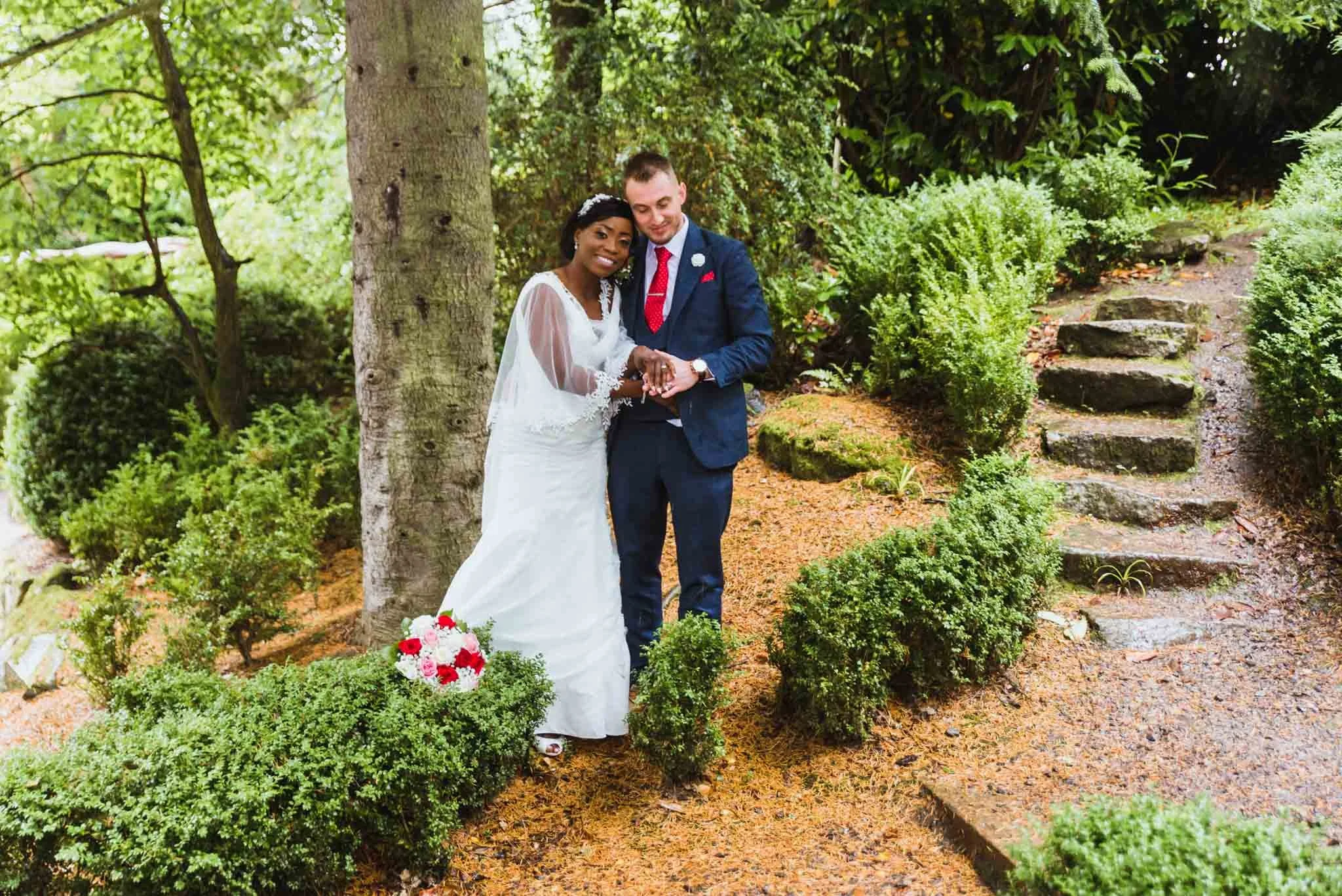 The couple hold eachother side by side beside a tree with stone steps to their right in a park for portraits.