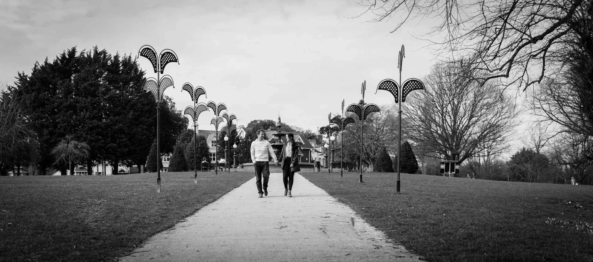 Mono image of the couple walking along a path with a fountain in the background.