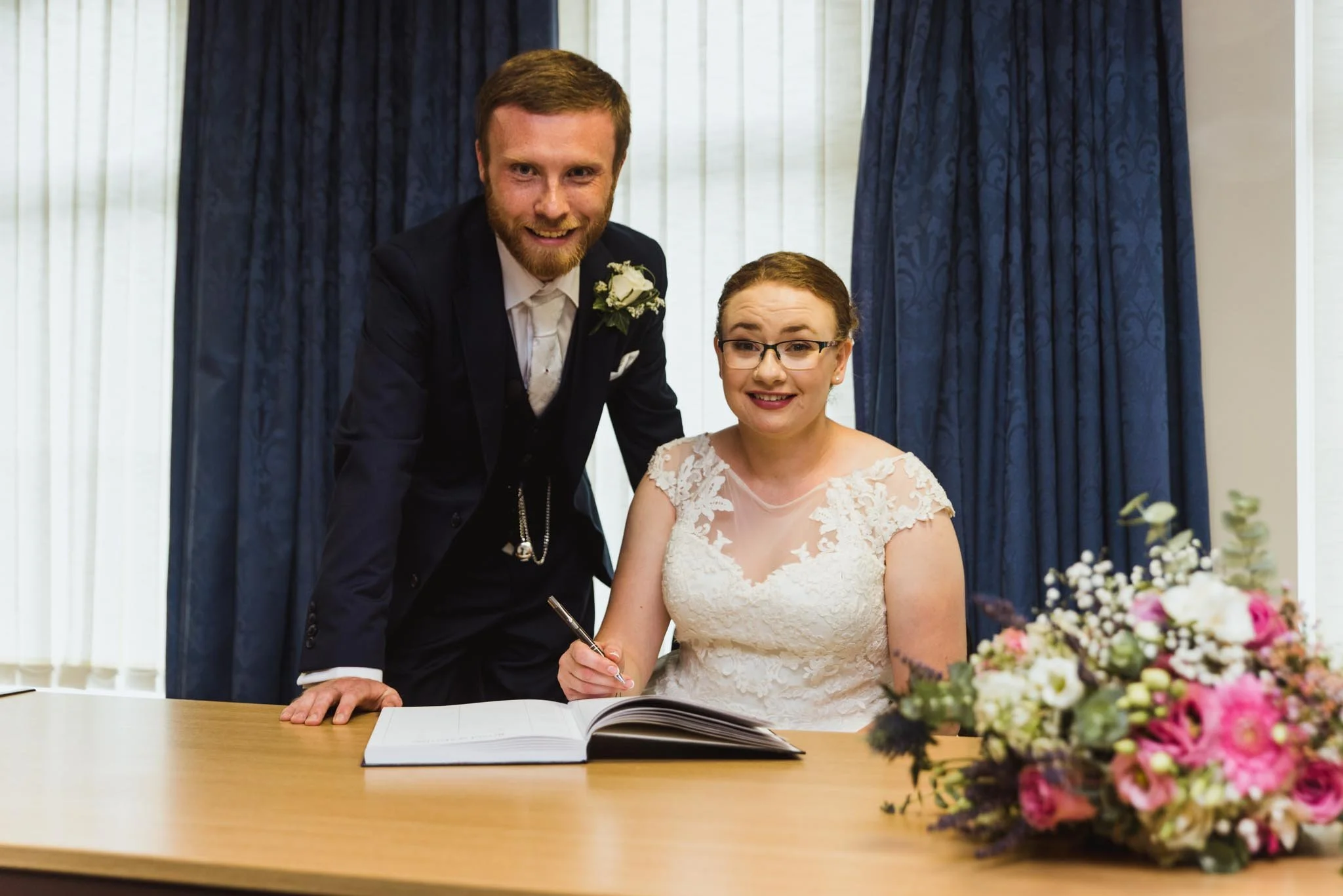 The bride and groom are signing the book with the bouquet on the table.